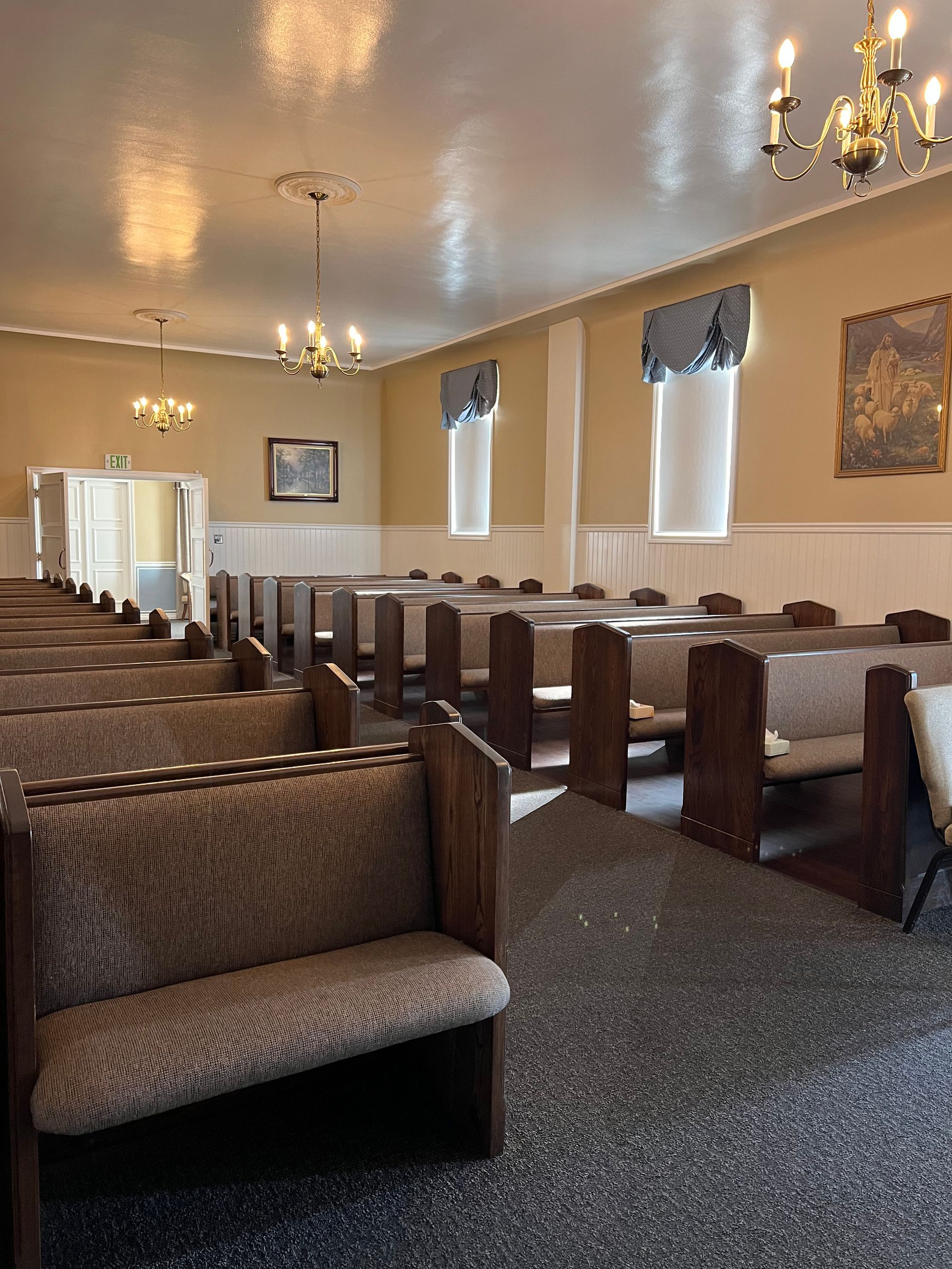 Empty church pews in a beige chapel with chandeliers, windows, and a patterned carpet.