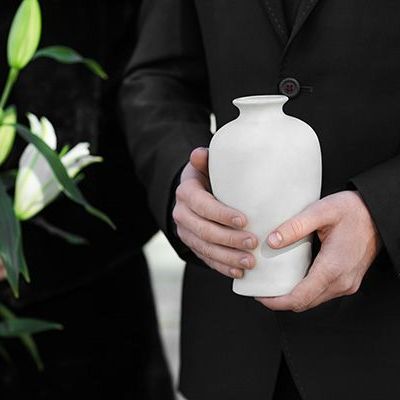 Hands in a black suit holding a white vase with greenery nearby