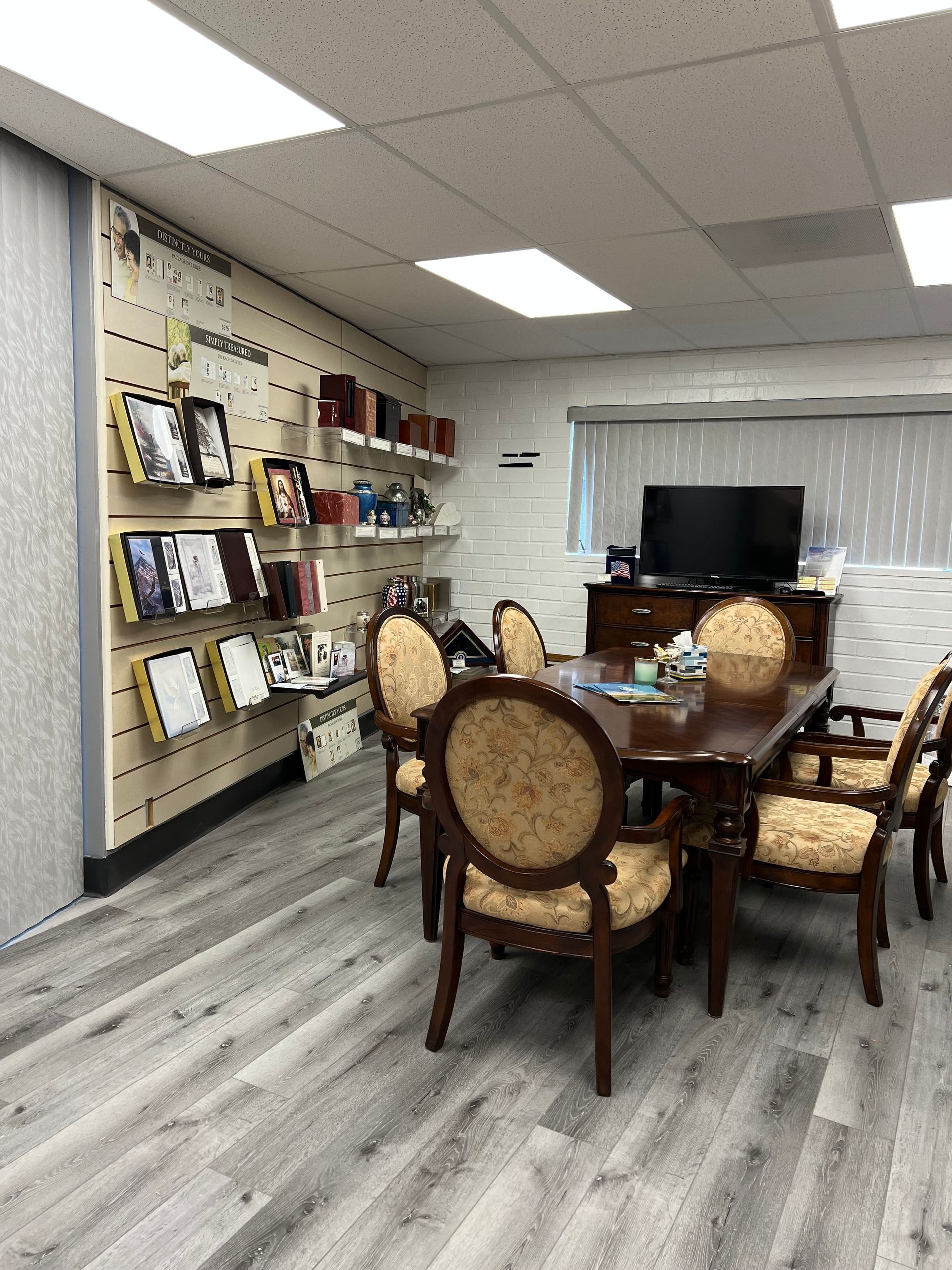 Office showroom with a conference table, chairs, wall displays, and a TV on a white brick wall.