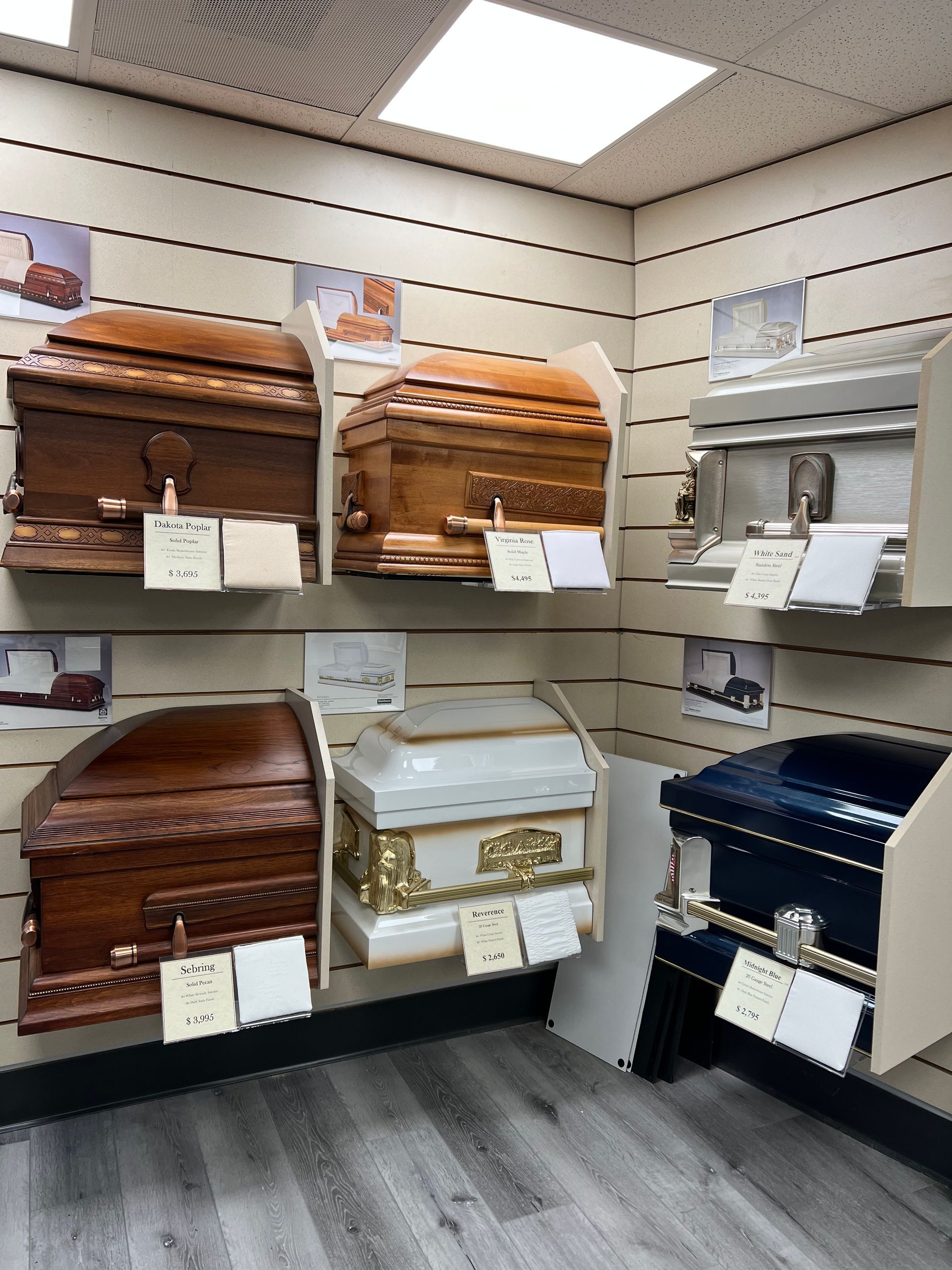 Caskets displayed in a showroom, including wood, white, and dark blue models on shelves.
