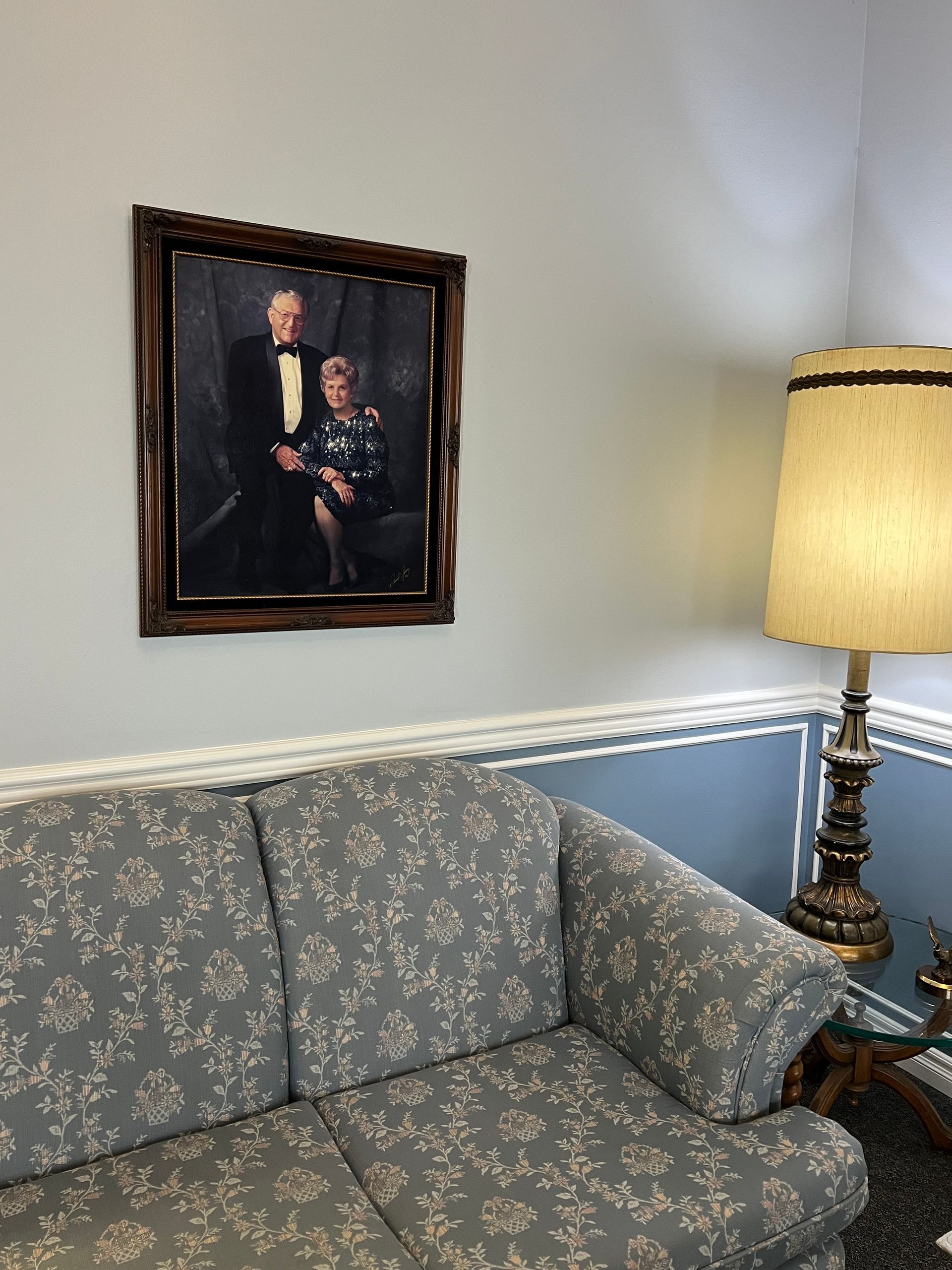 Floral loveseat beneath a framed portrait, beside a lit floor lamp in a softly lit room