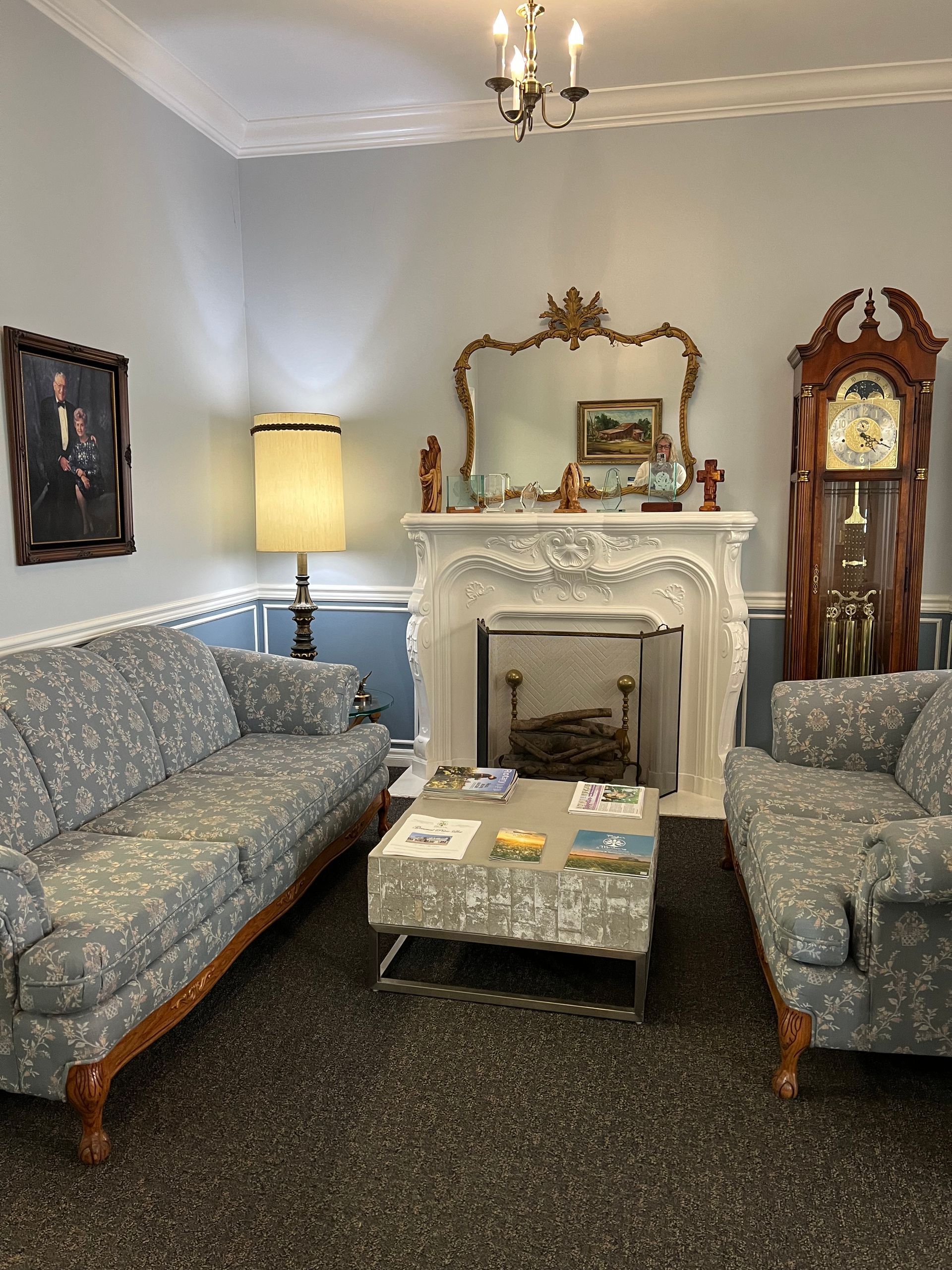 Cozy living room with gray sofas, ornate fireplace, tall clock, and lamp on carpeted floor