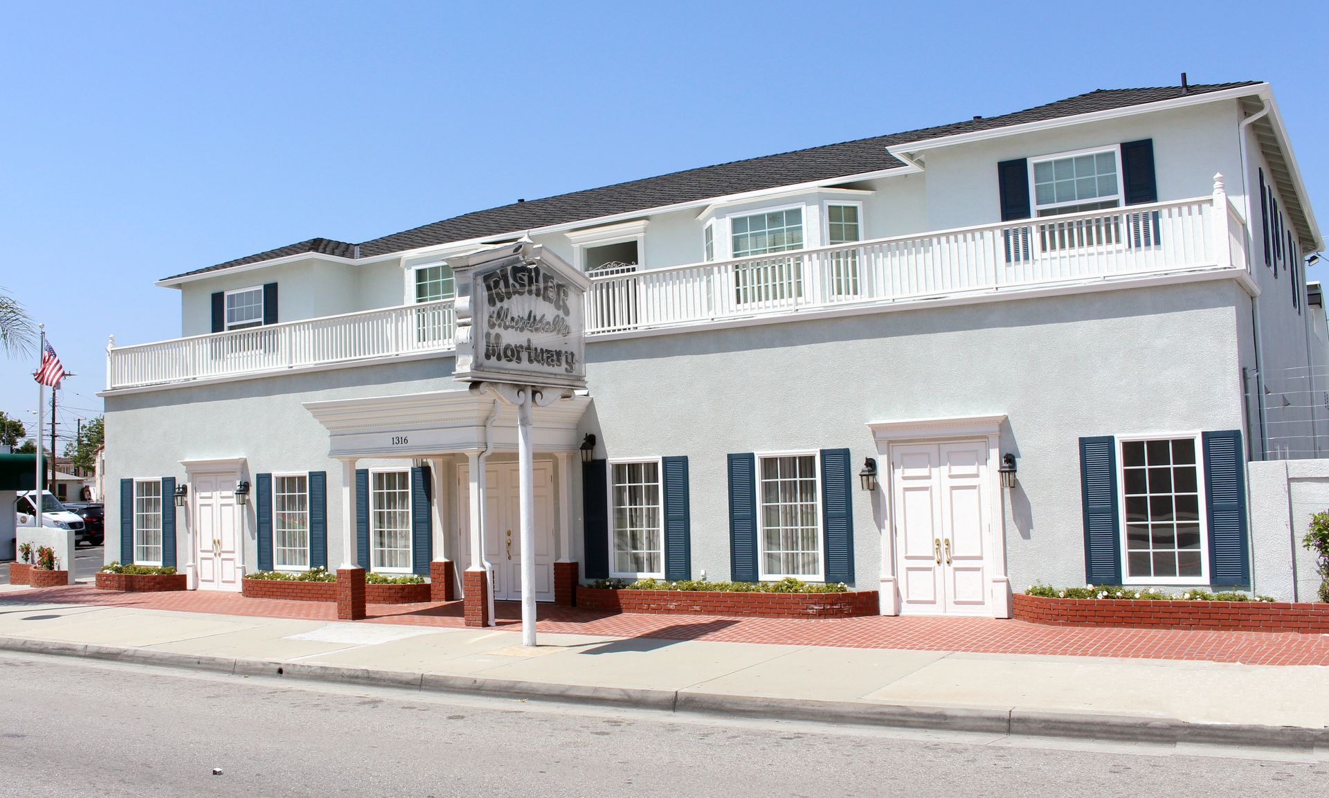 White two-story building with blue shutters and red flower boxes along a sunny street