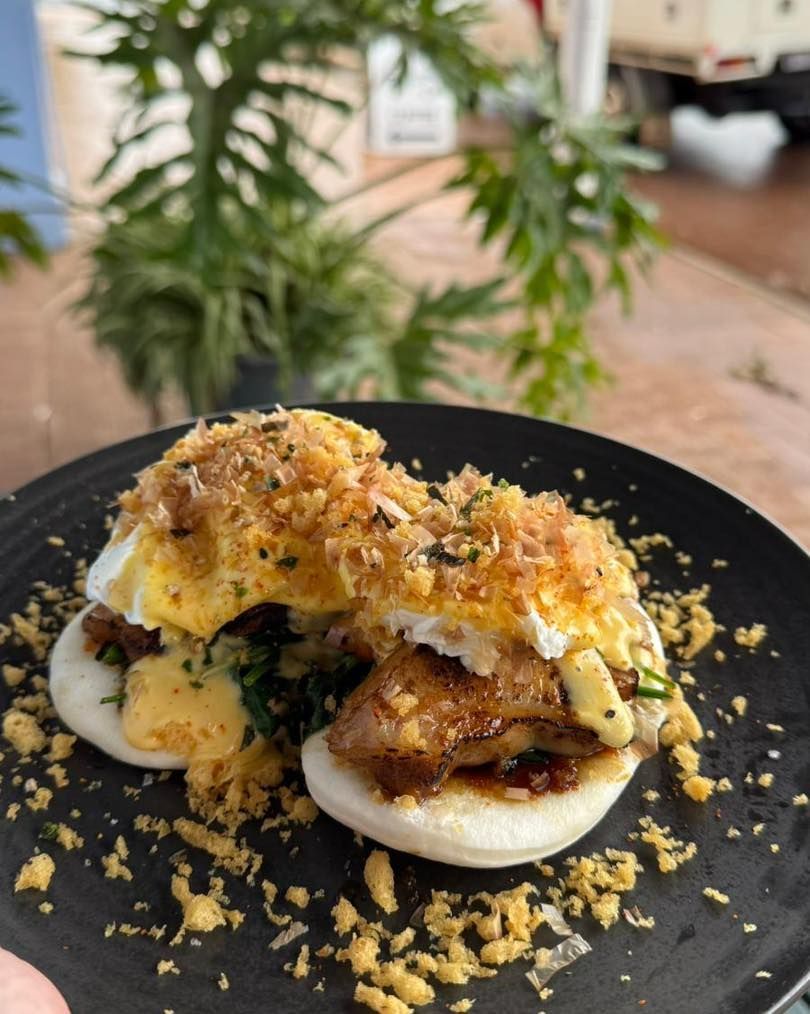 Two bao buns topped with egg, pork belly, and breadcrumbs on a black plate, outdoors.
