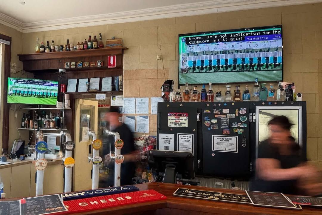 Bar interior with two TVs showing sports, a bartender, and a customer.