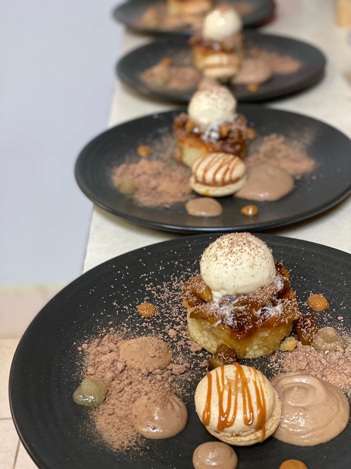Dessert plates: cake, ice cream, macaron, sauce, and cocoa powder. Dark plates arranged in a row.
