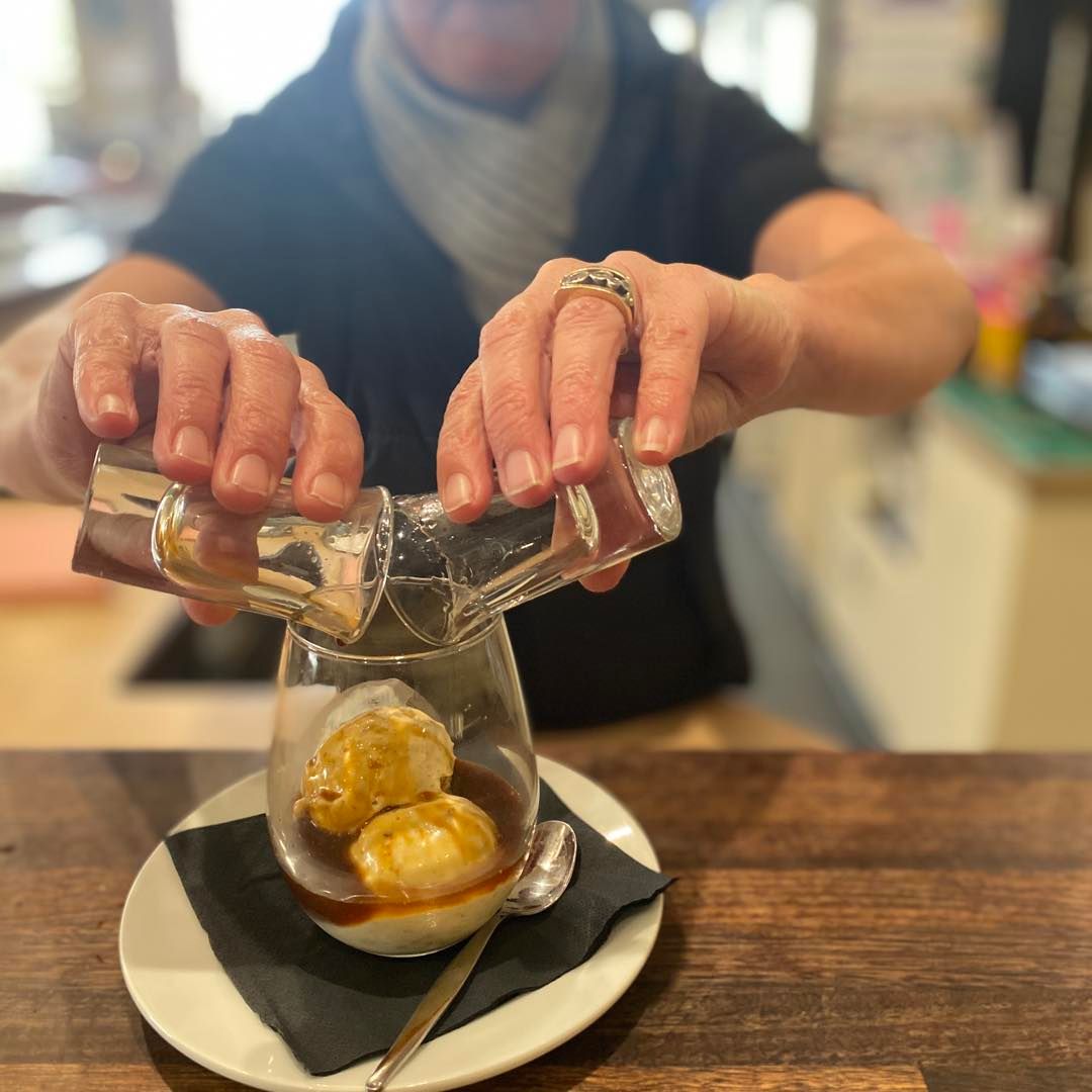 Person pouring liquid over ice cream in a glass, on a small plate atop a wooden bar.