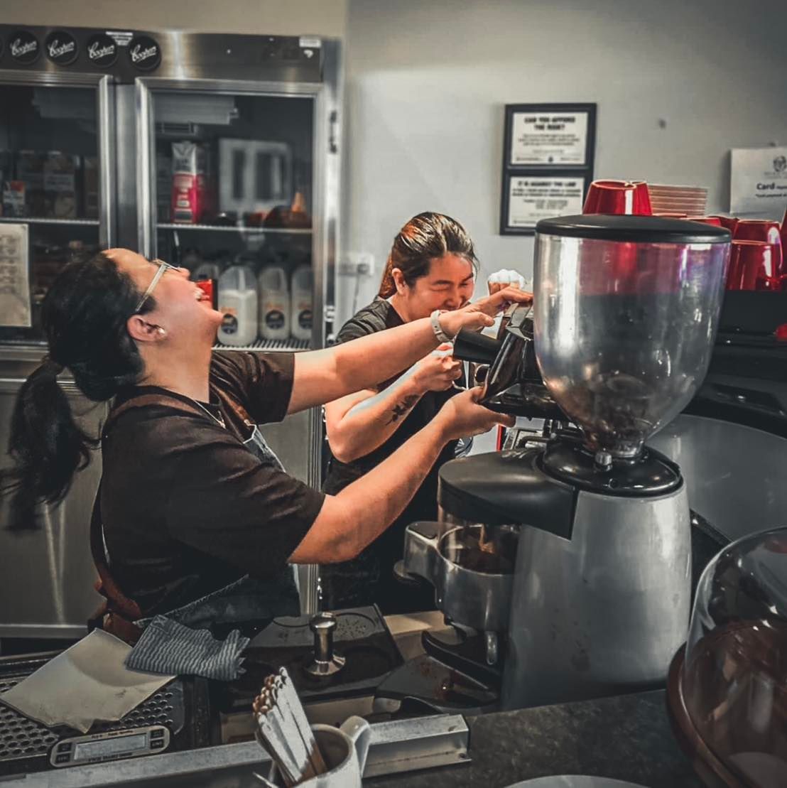 Two baristas laugh while working with a coffee grinder in a cafe.