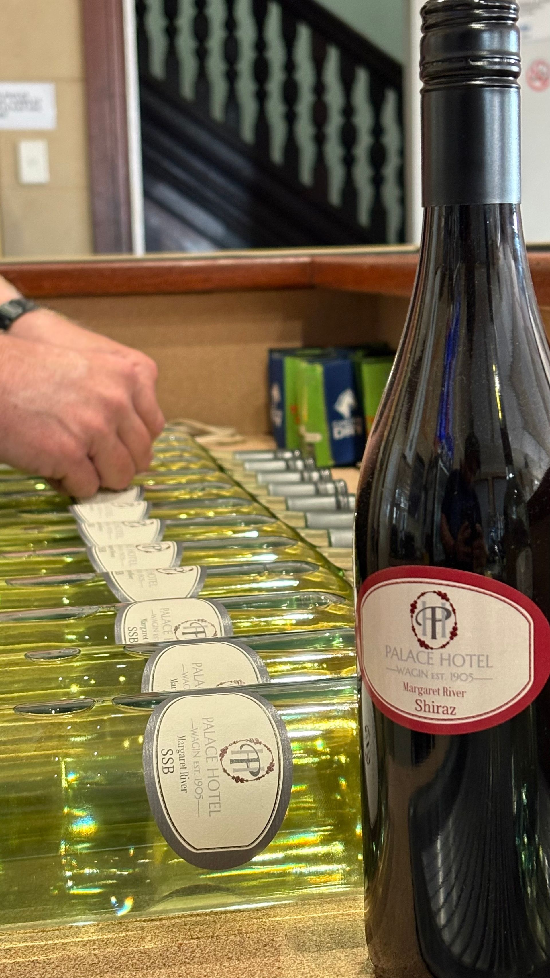 A hand reaching for a white wine bottle in a line of similar bottles. A dark wine bottle in focus to the right.