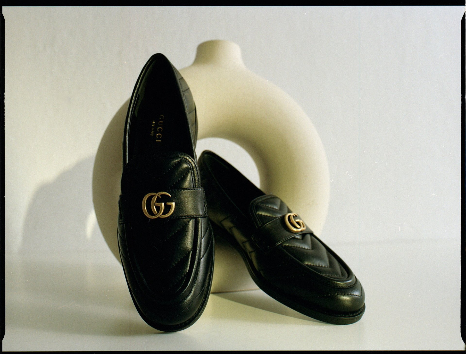 Schwarze Gucci-Loafer mit goldenem „GG“-Logo, an einer weißen Vase gelehnt.