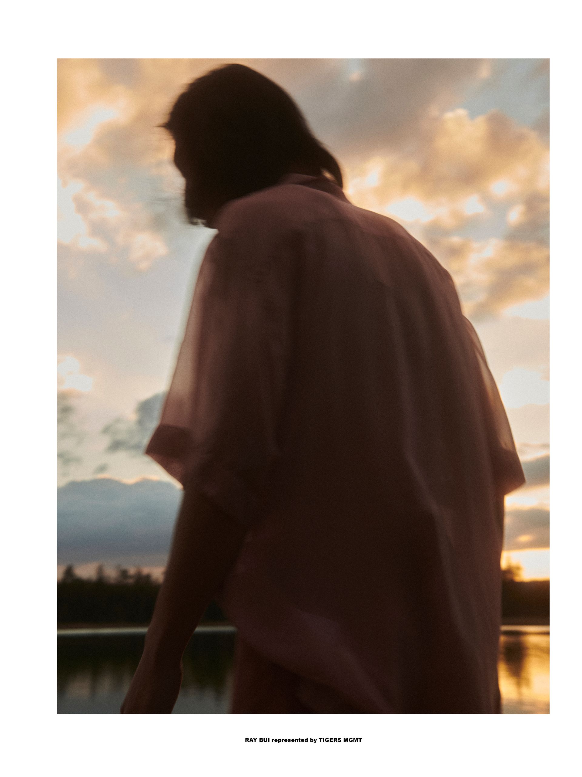 Person in einem rosa Hemd in der Abenddämmerung, die sich vor einem See und einem farbenfrohen Sonnenuntergang abhebt.