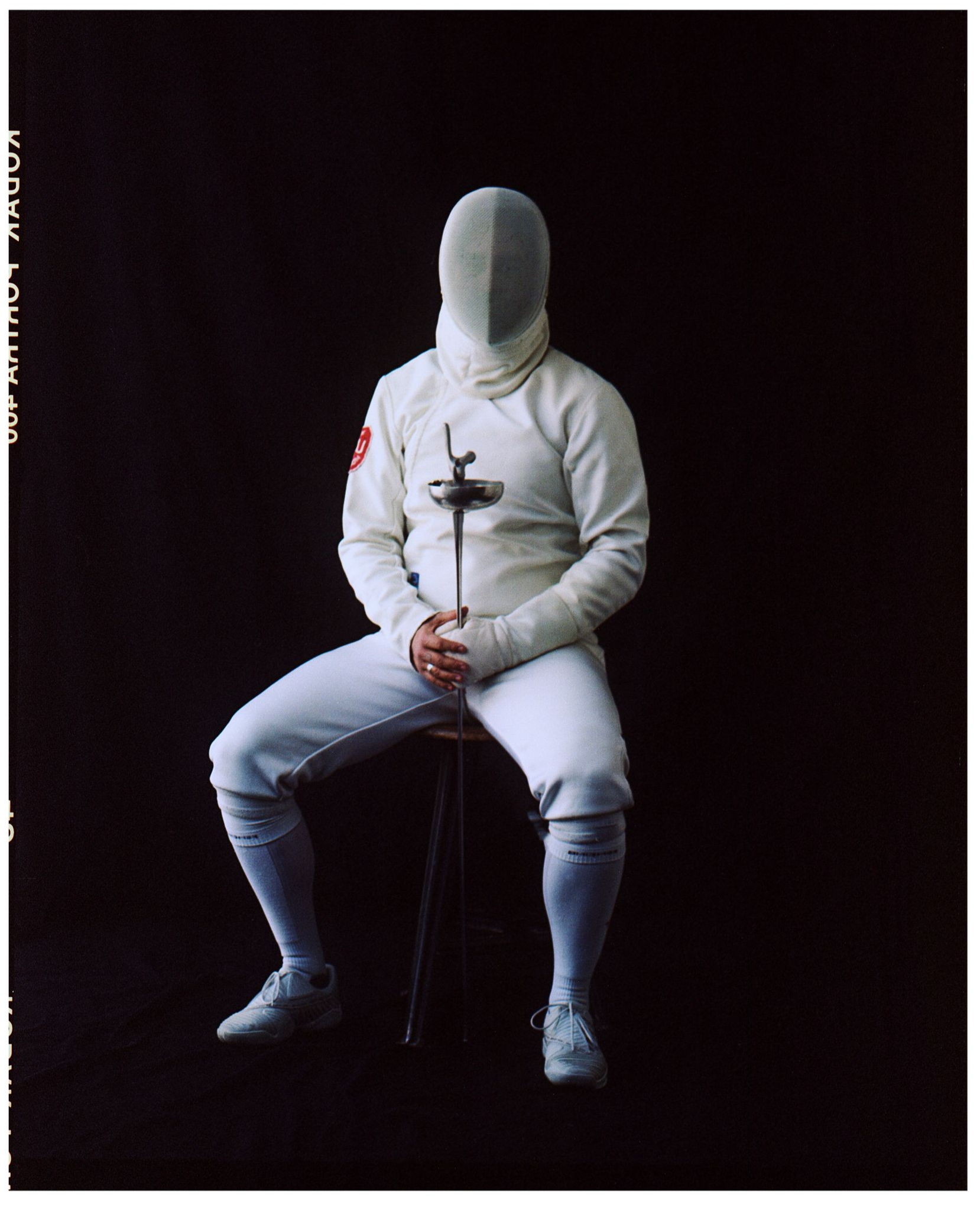 Fechter in weißer Uniform, Florett haltend, auf einem Hocker vor schwarzem Hintergrund sitzend.