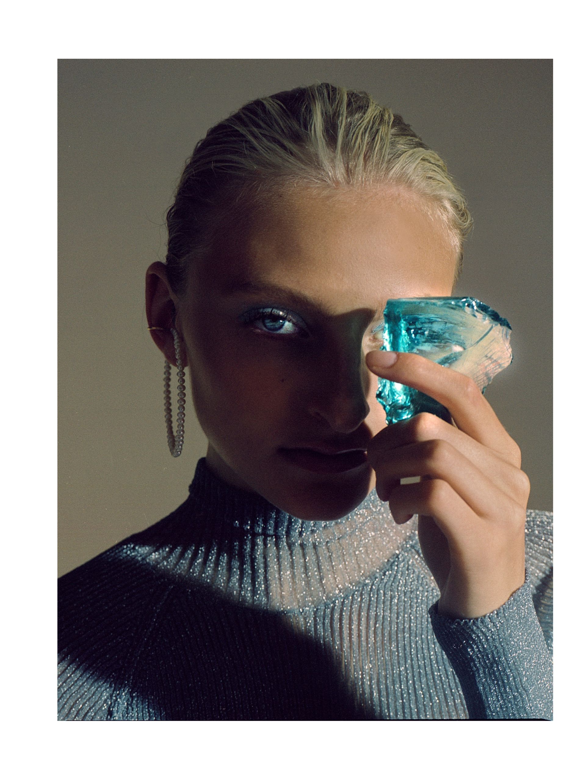 Frau, die einen blauen Kristall an ihr Auge hält und einen glitzernden Rollkragenpullover und Creolen trägt.