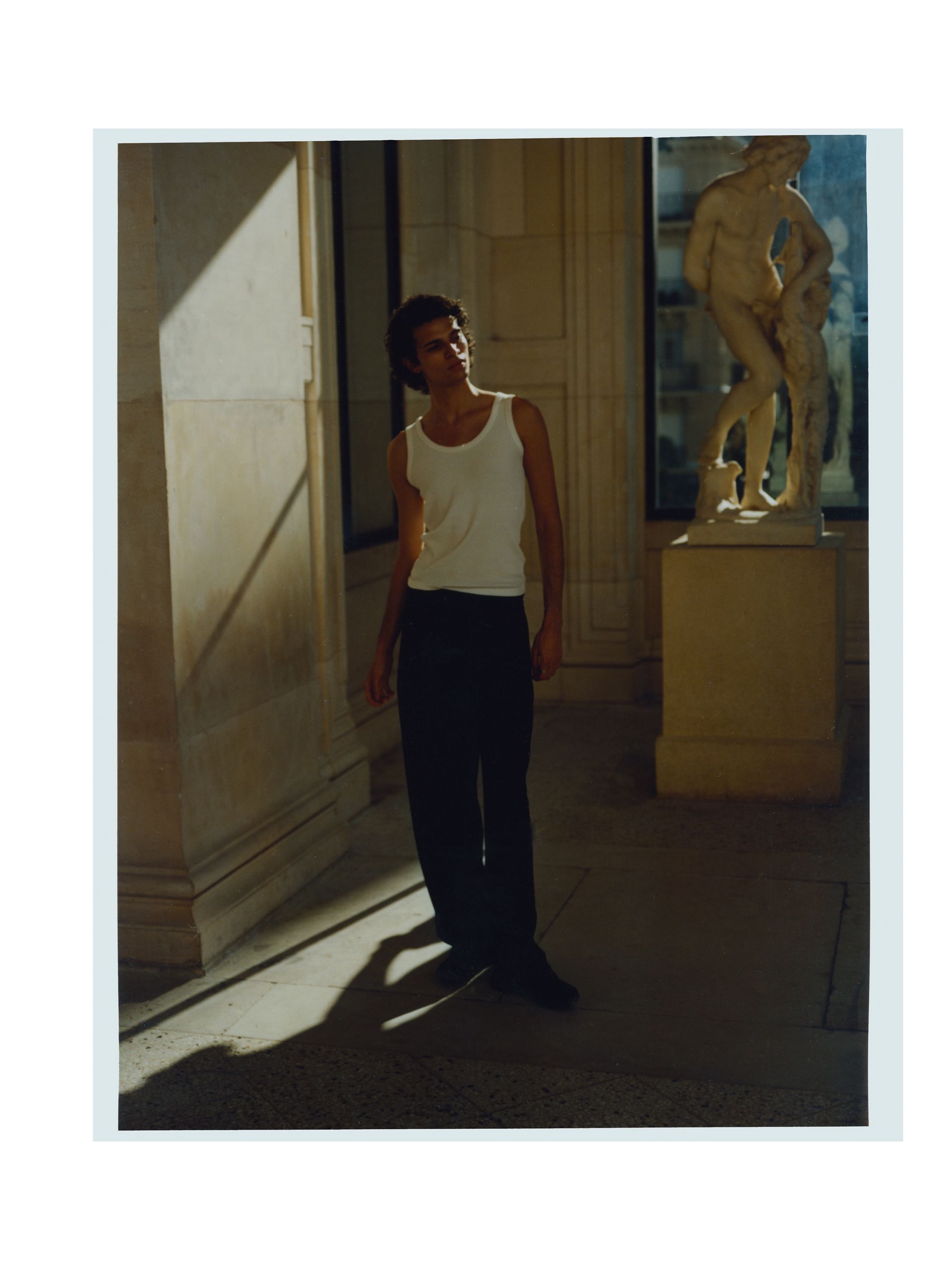 Ein Mann in einem weißen Tanktop und einer schwarzen Hose steht neben einer Statue in einem sonnendurchfluteten Museum.