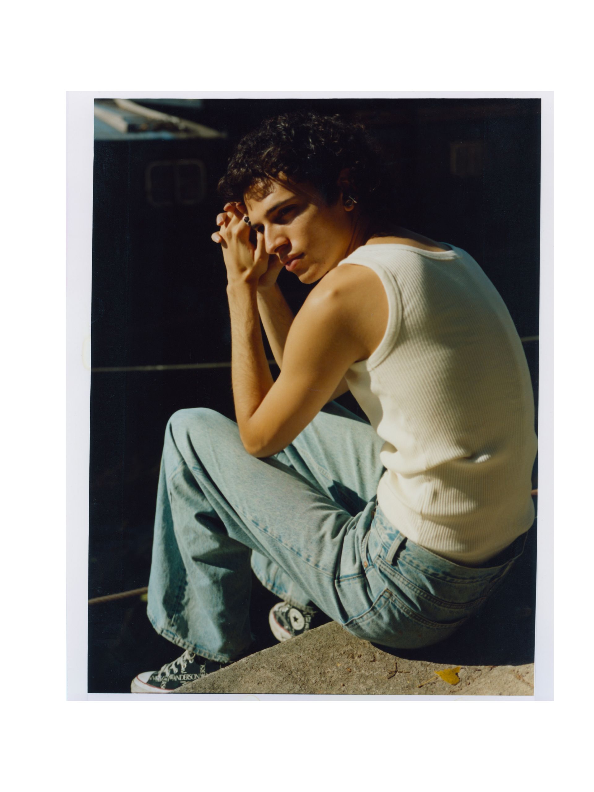 Sitzende Person in Jeans und Tanktop, Hände vor dem Gesicht, Blick nach unten, im Freien.