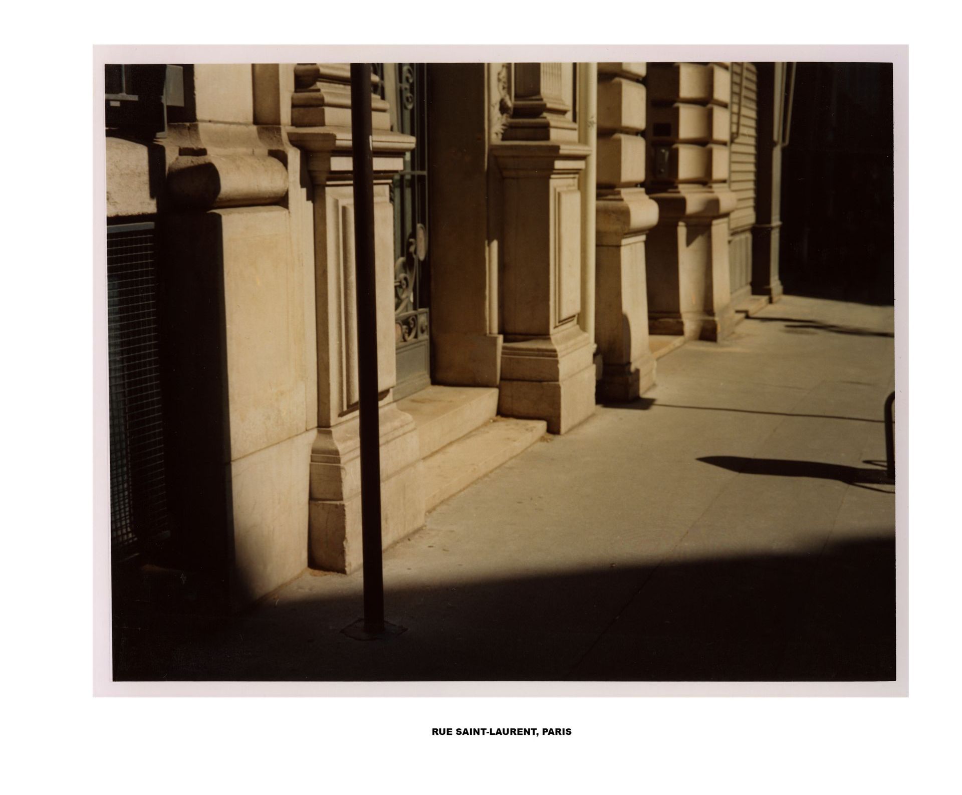 Gebäudefassade mit Säulen, die lange Schatten auf einen Bürgersteig werfen. Beige- und Brauntöne.