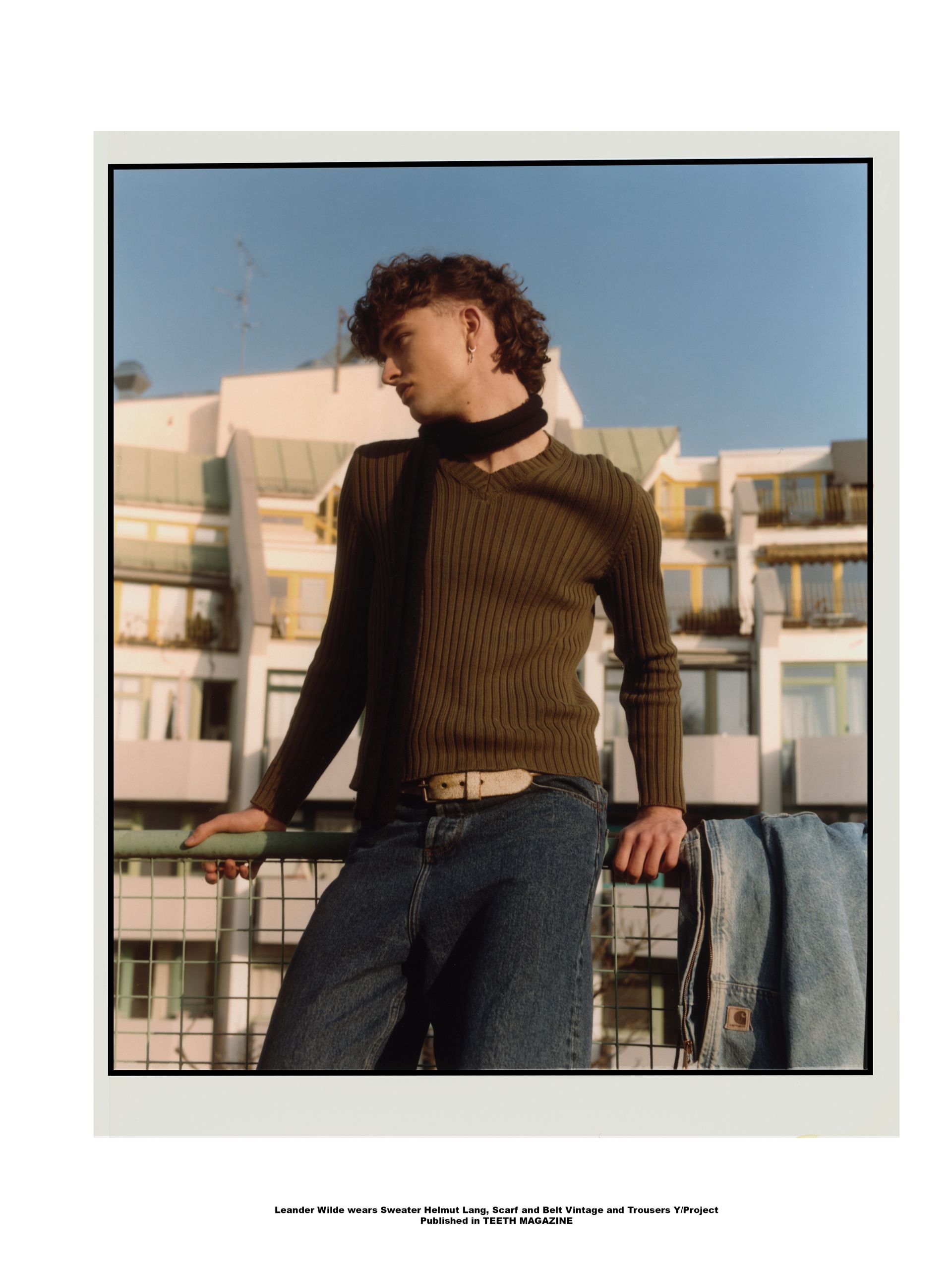 Mann mit lockigem Haar, bekleidet mit grünem Pullover, blauen Jeans und Schal, steht auf einem Balkon.