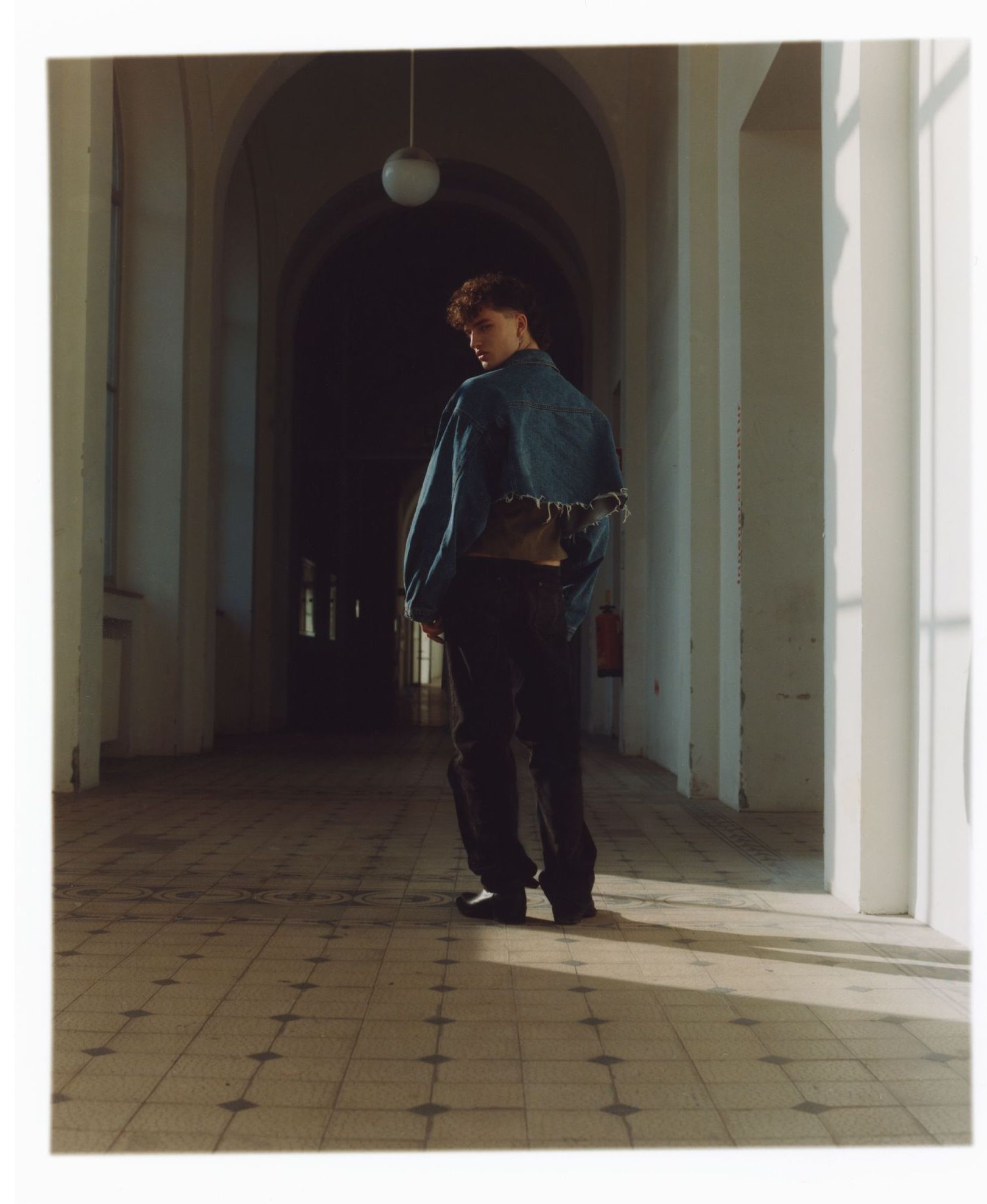 Eine Person in Jeansjacke und dunkler Hose steht mit dem Rücken zur Kamera in einem langen, gewölbten Flur. 