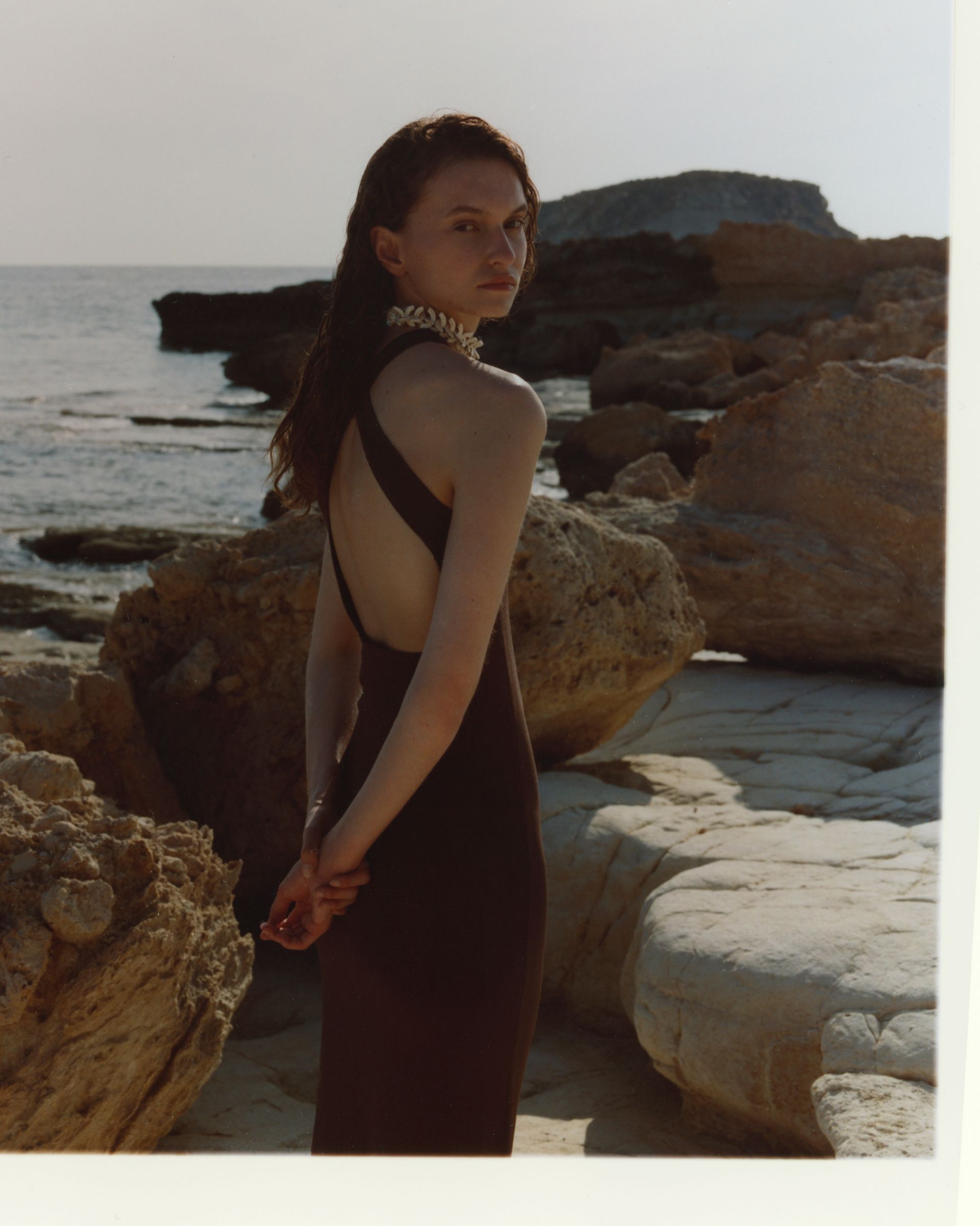 Frau im rückenfreien braunen Kleid steht am felsigen Strand und blickt über ihre Schulter.