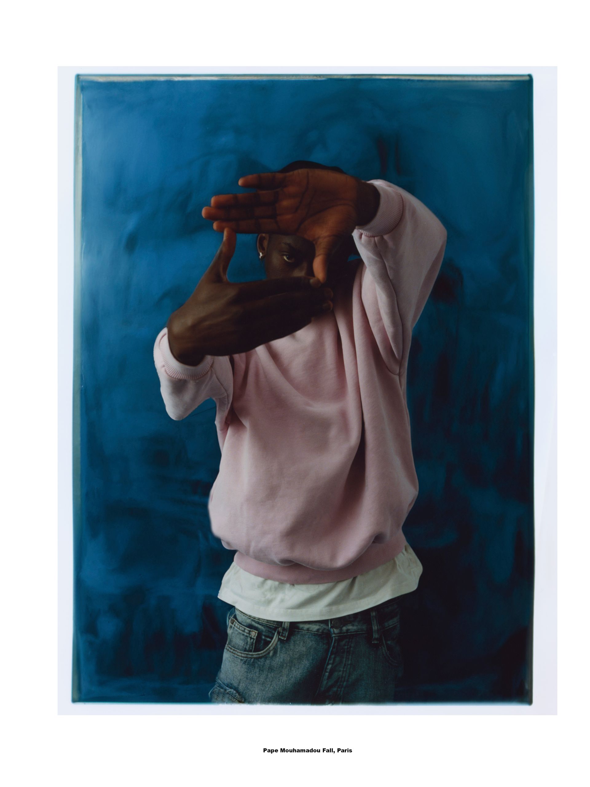 Person umrahmt Gesicht mit Händen; blauer Hintergrund; trägt rosa Sweatshirt und Jeans.