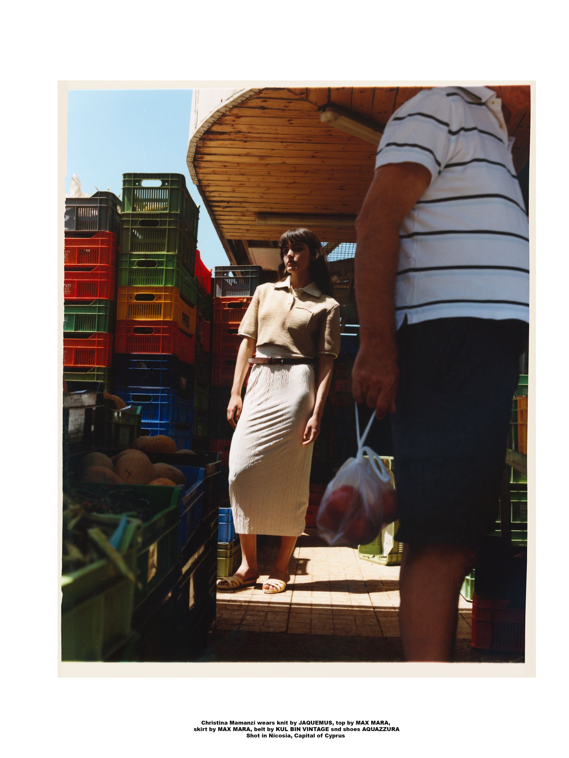 Frau in beigem Outfit steht neben bunten Kisten auf einem Markt, ein Mann hält eine Tüte mit Produkten.