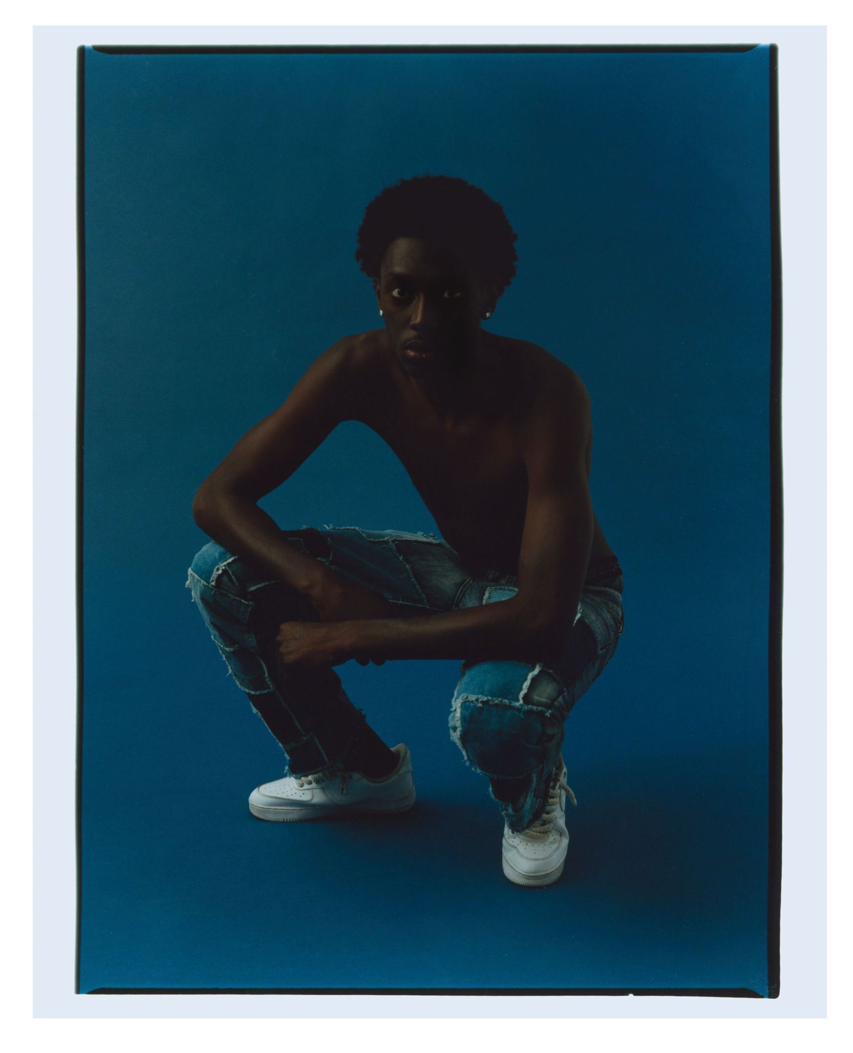 Eine Person ohne Hemd hockt auf einem blauen Hintergrund und trägt zerrissene Jeans und weiße Turnschuhe.