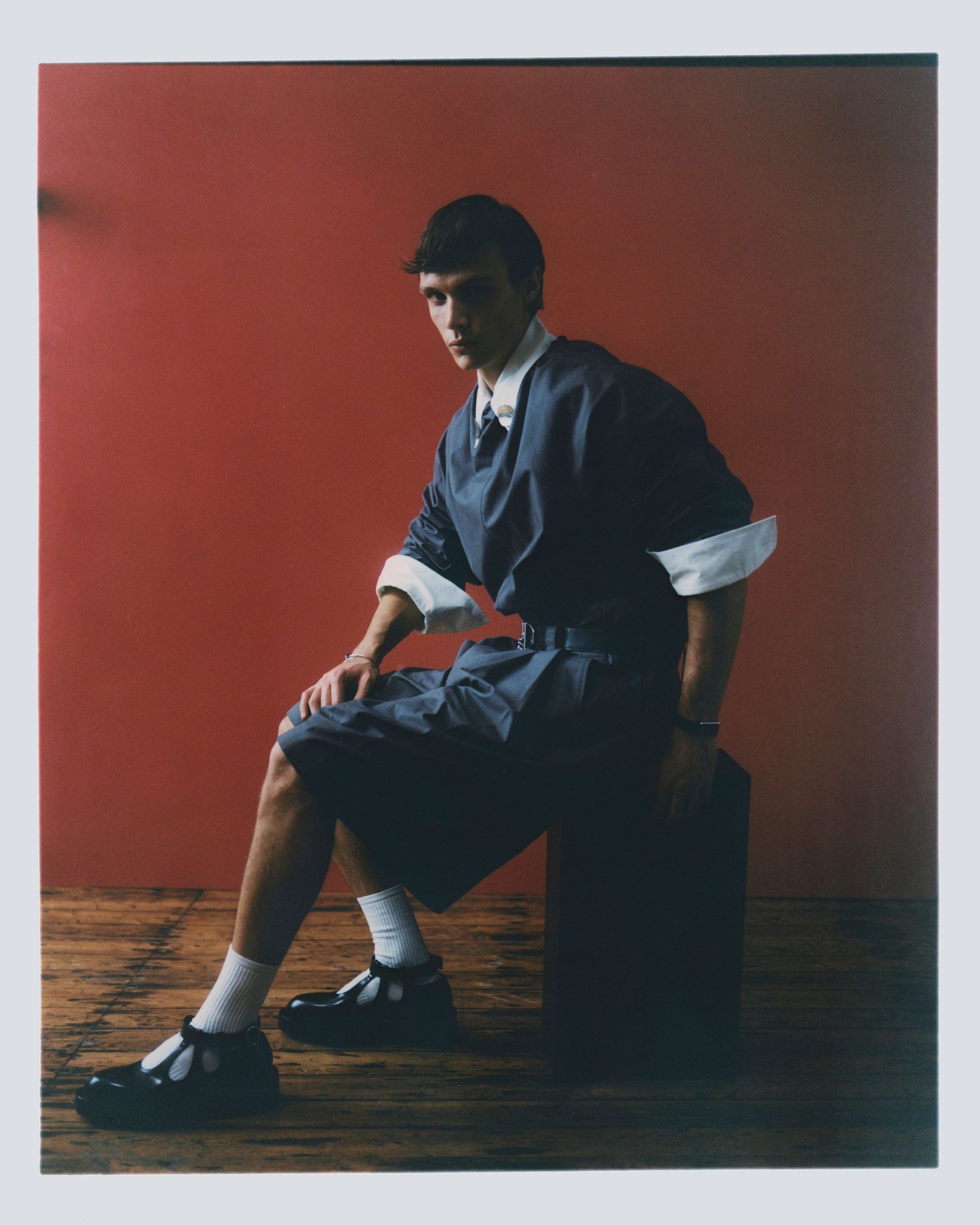 Mann in blauem Hemd und Shorts, weißen Socken, schwarzen Schuhen, sitzt auf einem Block vor einer roten Wand.