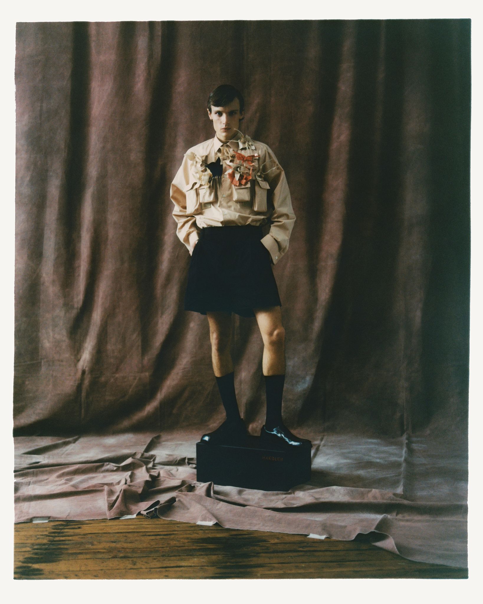 Person in Kilt, Stiefeln und hellbraunem Hemd steht auf einer Kiste. Hintergrund: brauner drapierter Stoff.