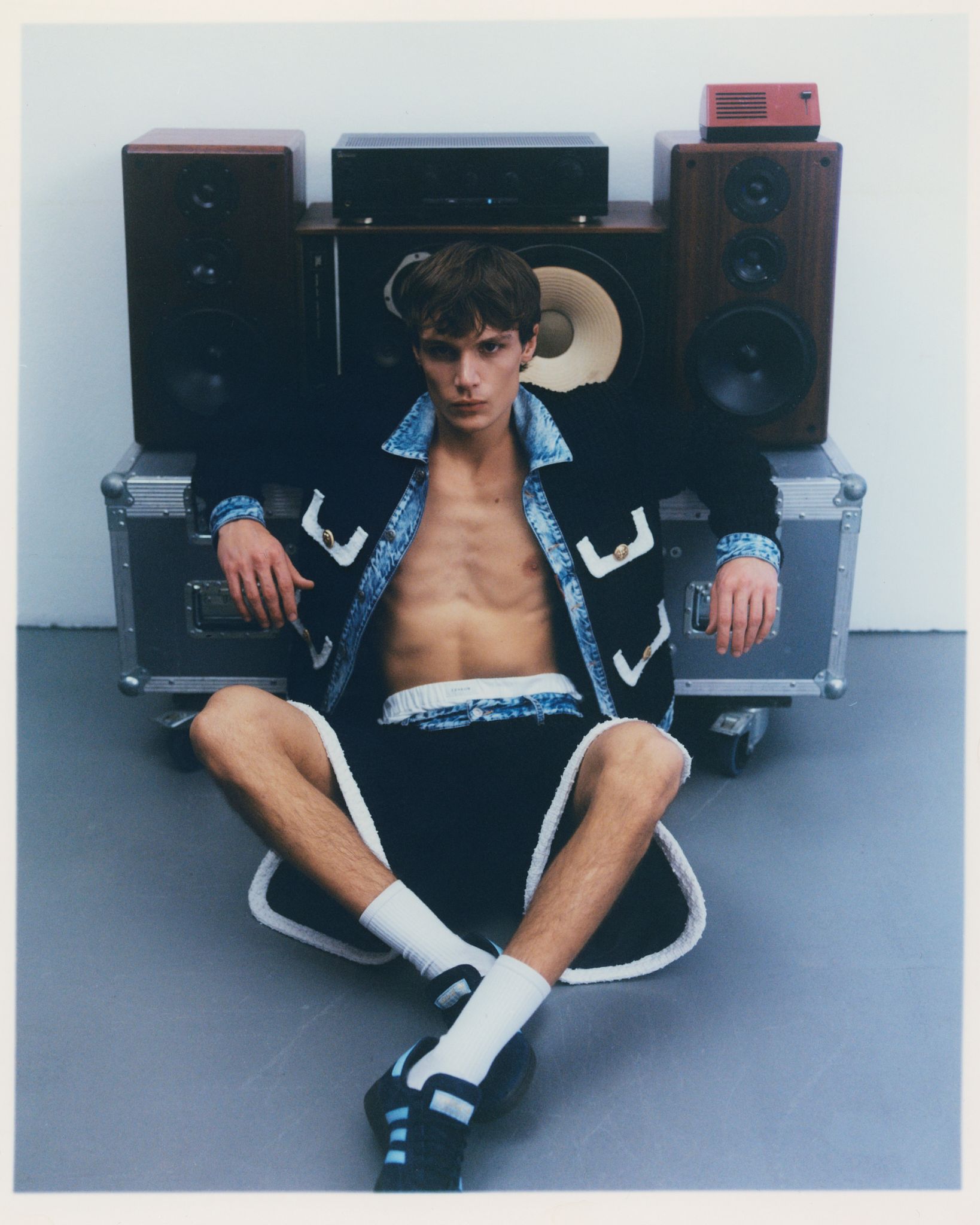 Mann in Jeansjacke und Shorts sitzt vor Lautsprechern. Grauer Raum, weiße Socken.