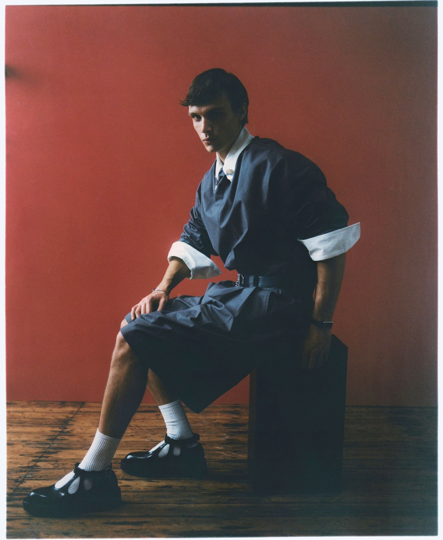 Mann in blauem Hemd und Shorts, weißen Socken, schwarzen Schuhen, sitzt auf einem Block vor einer roten Wand.