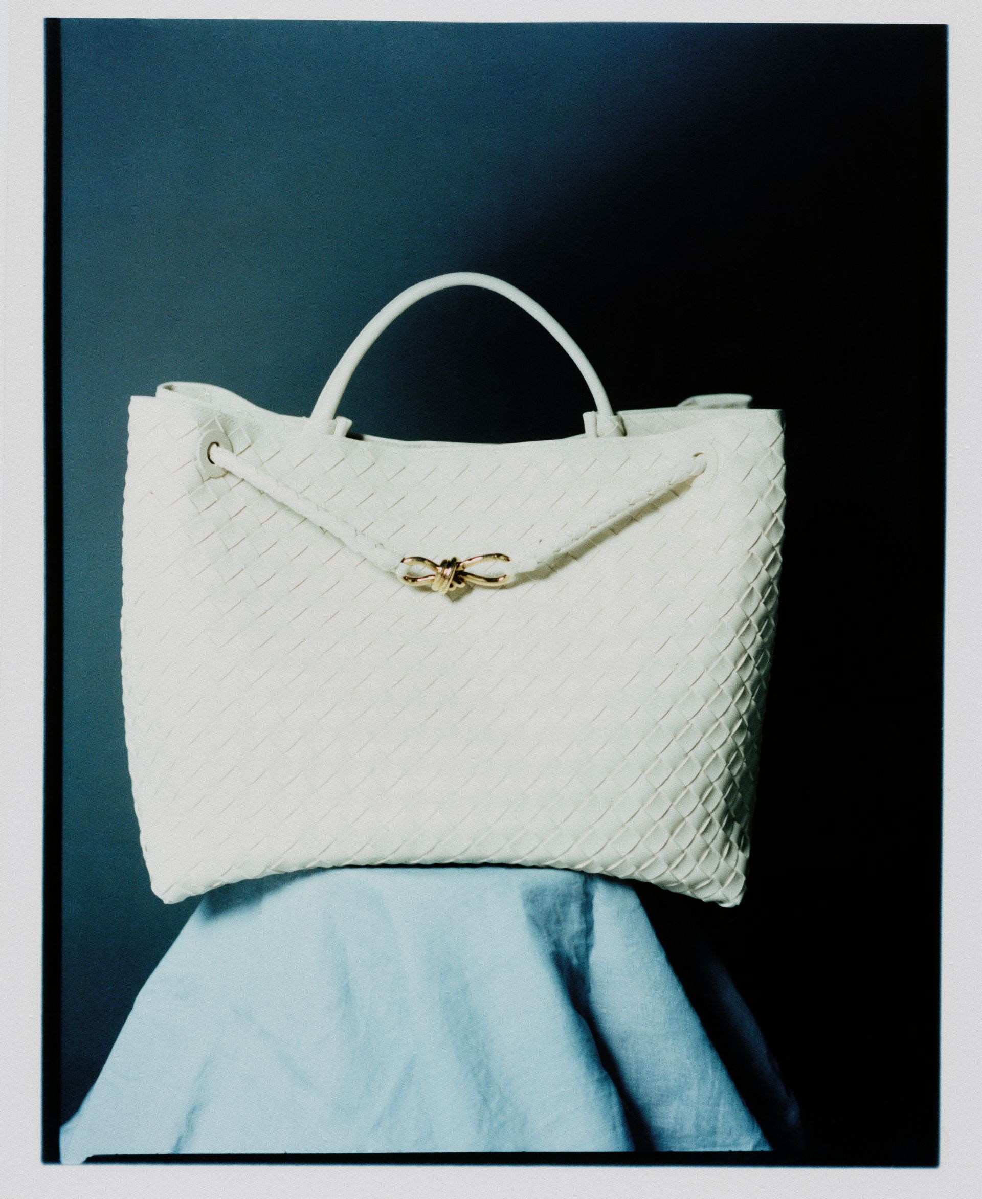 Weiße Handtasche auf blauem Stoff, vor dunklem Hintergrund.