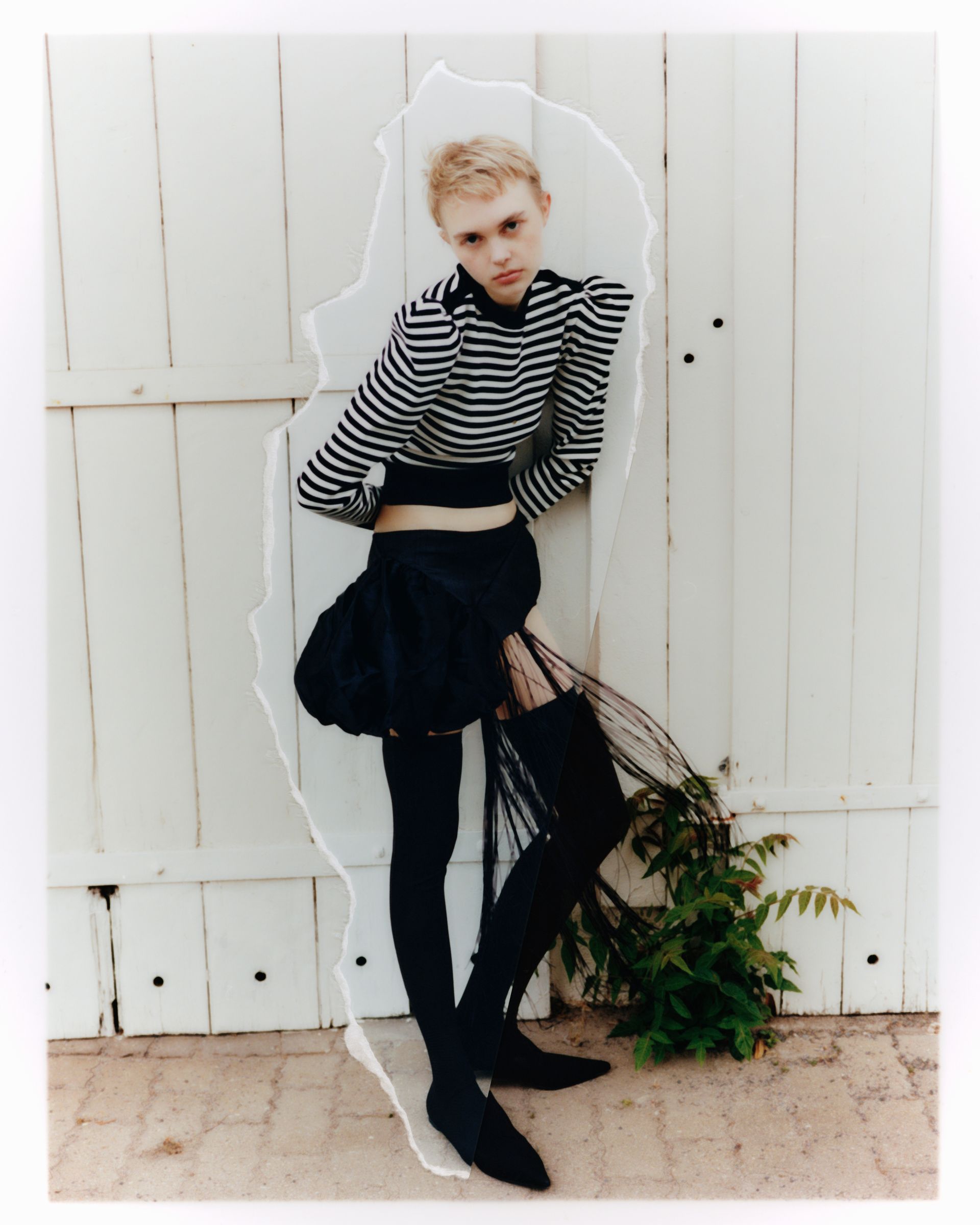 Person mit kurzen blonden Haaren, schwarz-weiß gestreiftem Hemd und schwarzem Rock, an weißer Tür gelehnt.