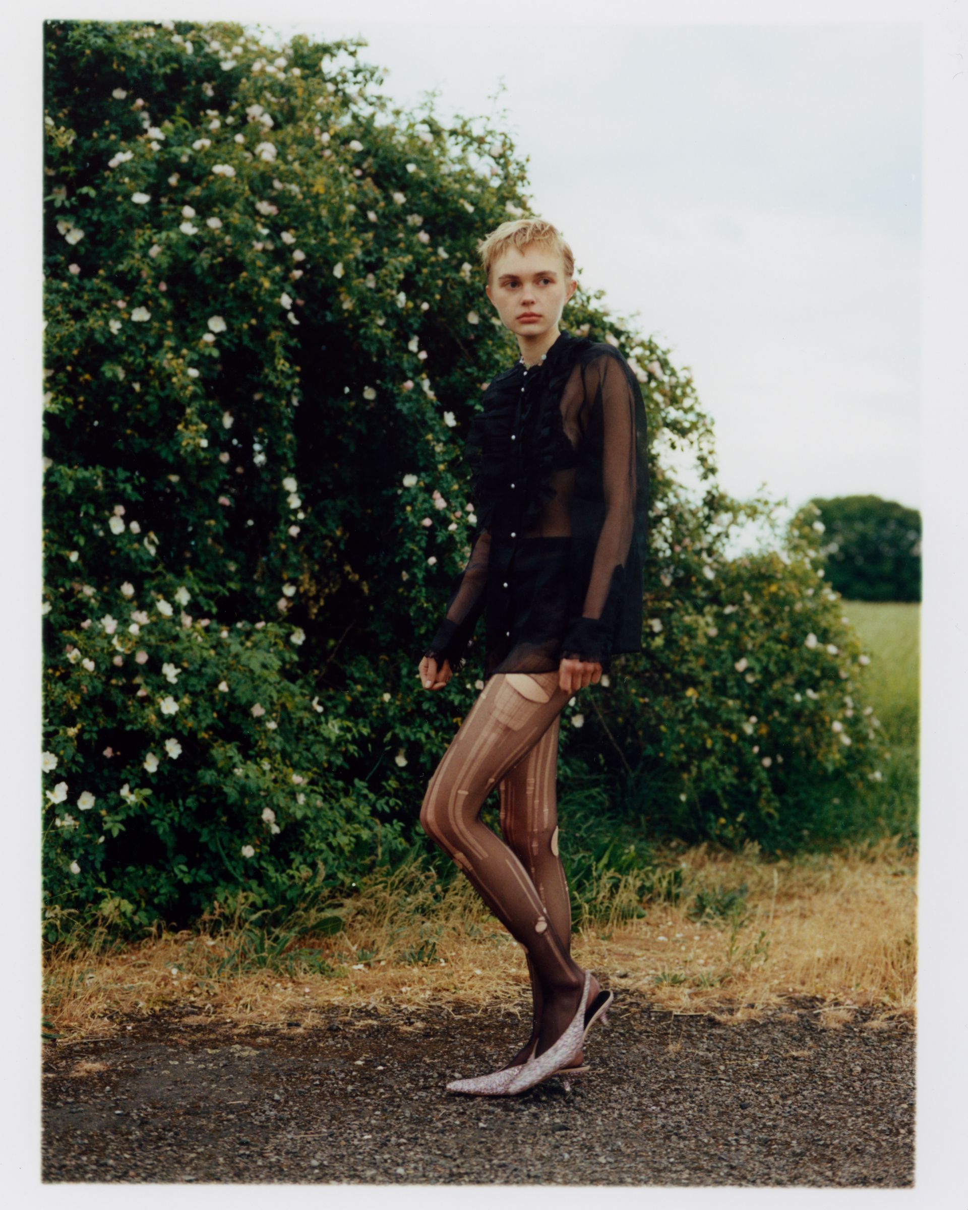 Mann in transparentem Hemd, Strümpfen und High Heels steht auf einem Weg. Grüner Busch im Hintergrund, ländliche Umgebung.