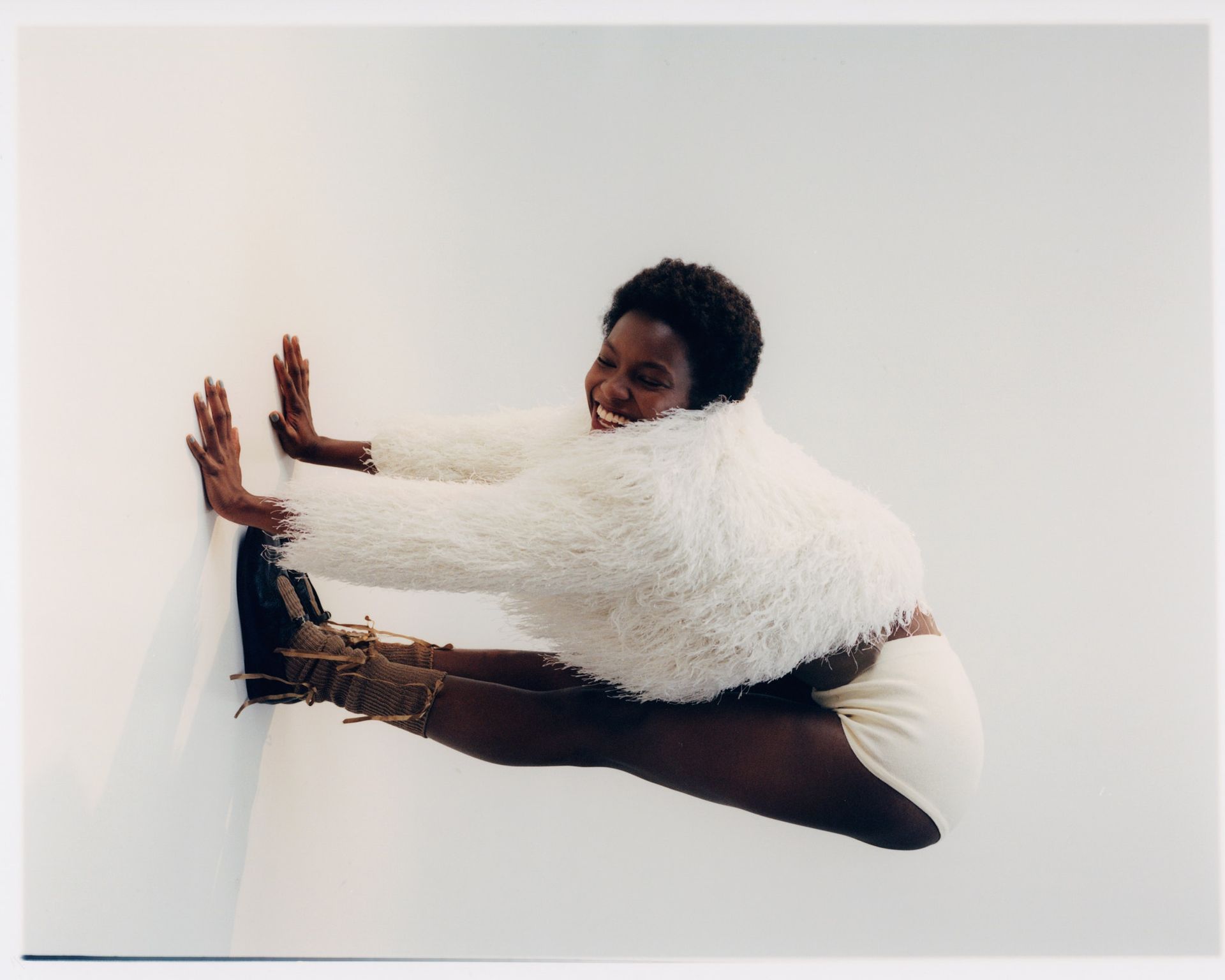 Frau in weißem Flauschpullover und Shorts lächelt und streckt ihre Beine vor einer weißen Wand aus.