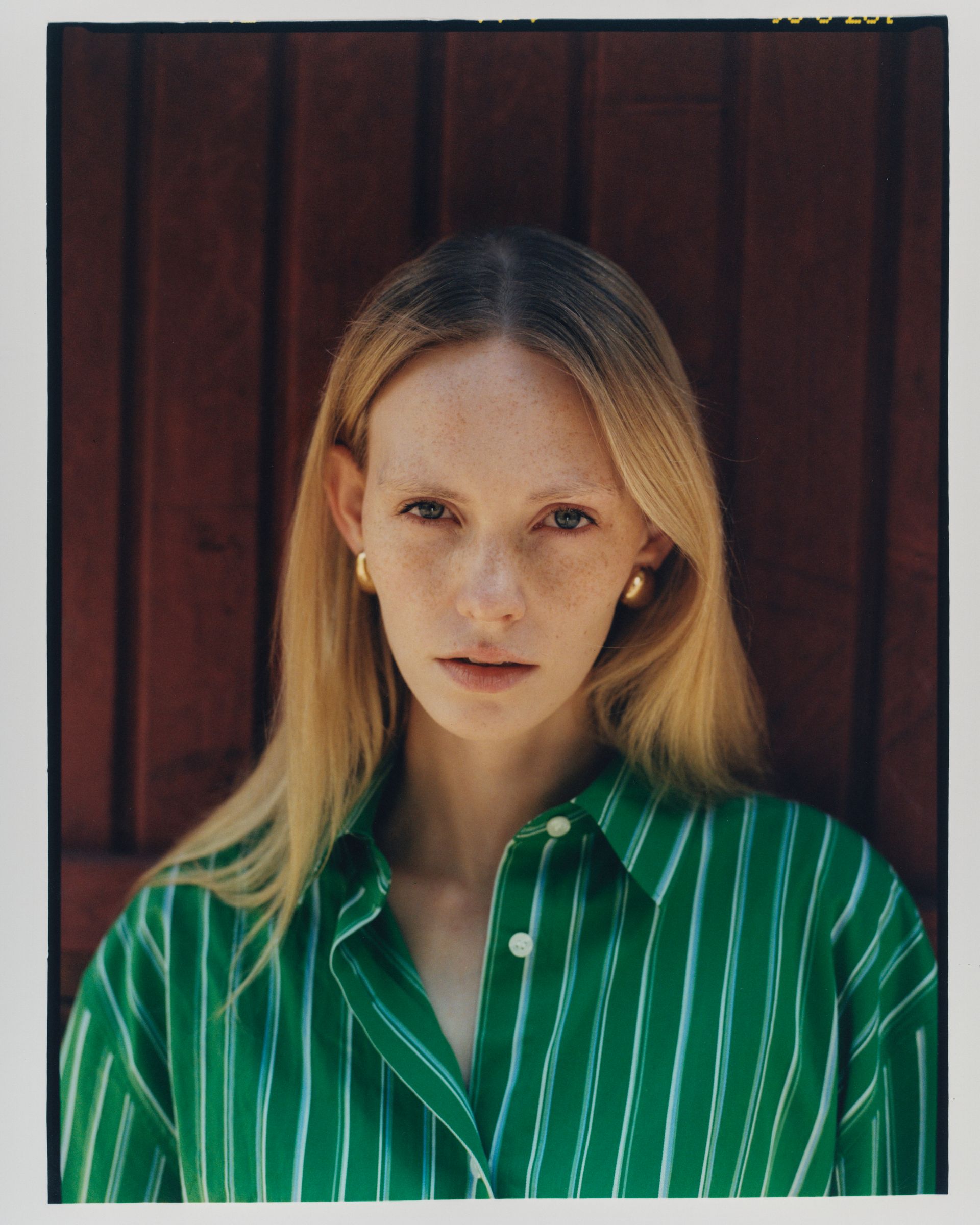 Frau mit blonden Haaren und Sommersprossen, trägt ein grün gestreiftes Hemd, blickt in die Kamera, vor einer roten Holzwand.
