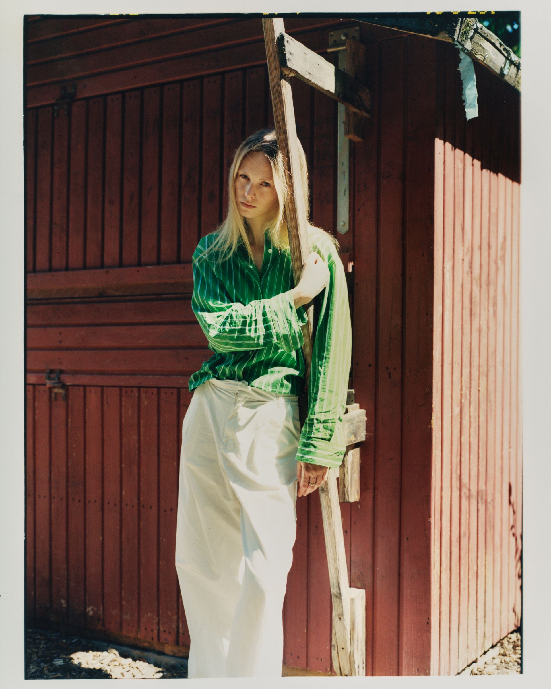 Frau in grünem Hemd und weißem Rock lehnt im Sonnenlicht an einem roten Holzschuppen.