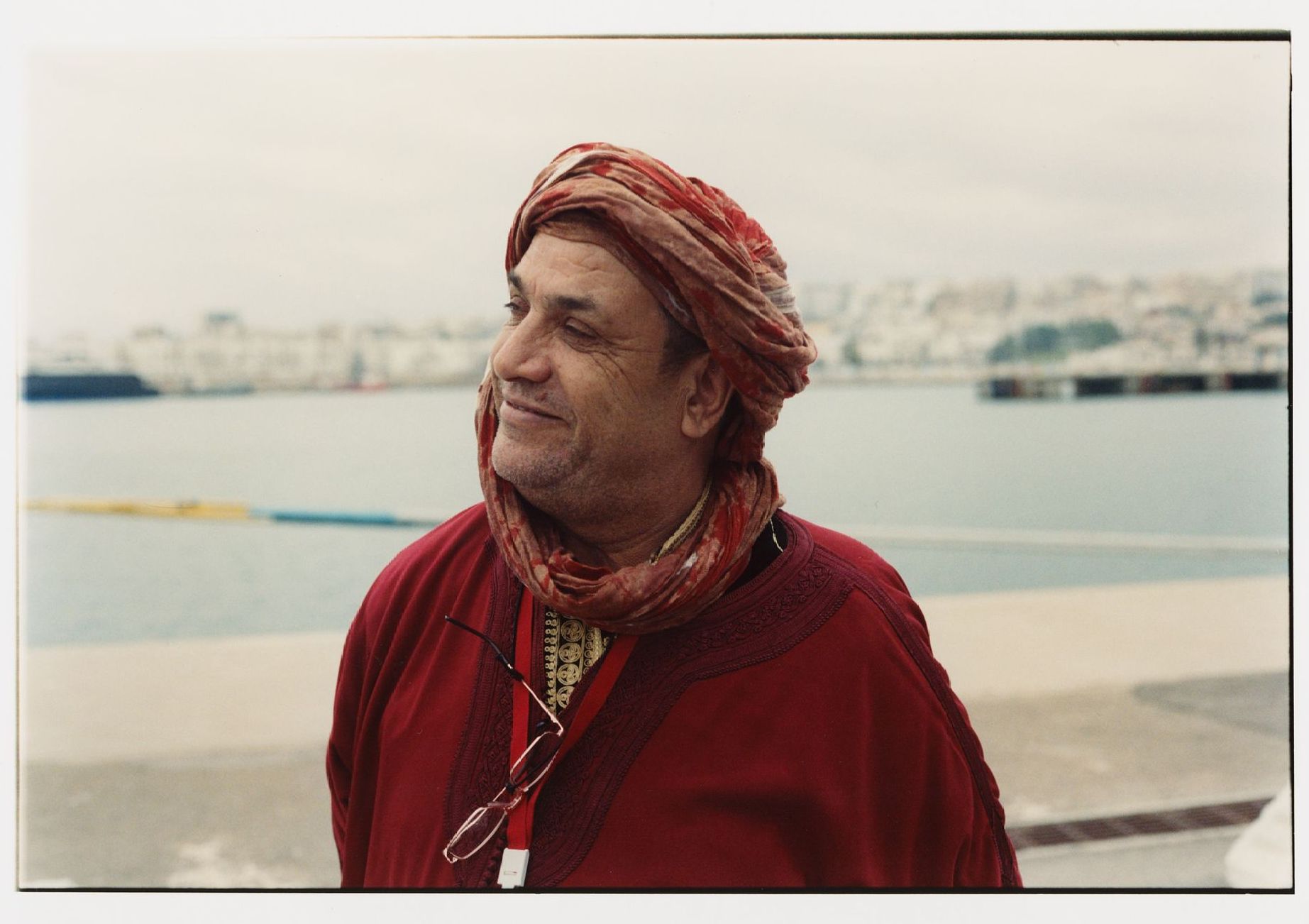 Mann mit rotem Turban und Robe am Wasser, blickt weg und lächelt. Stadt im Hintergrund.