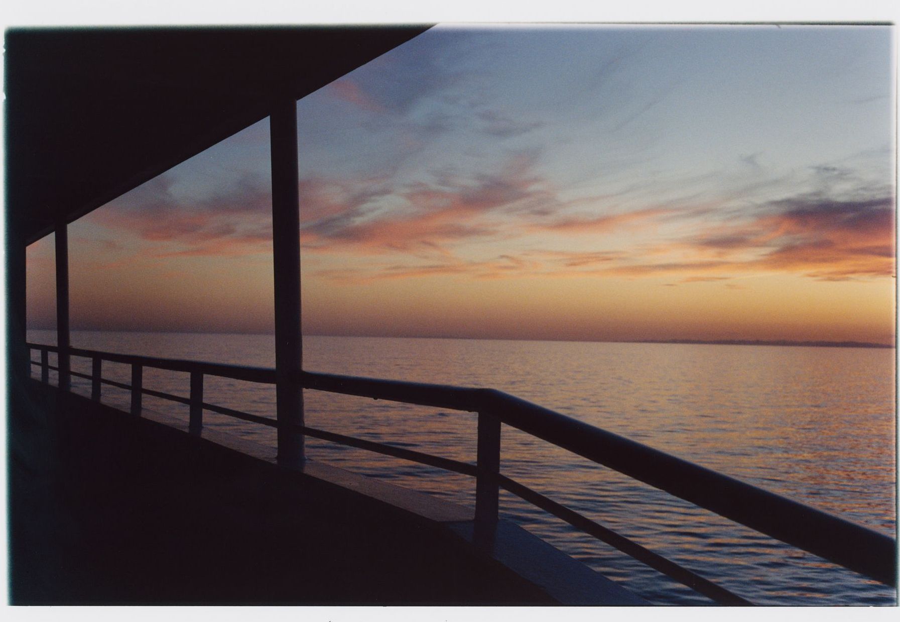 Sonnenuntergang über ruhigem Wasser, von einem Deck mit Geländer aus betrachtet, als Silhouette vor dem farbenfrohen Himmel.
