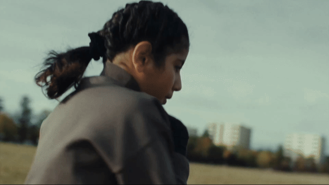 Mädchen mit zurückgebundenem dunklem Haar blickt auf eine Wiese hinunter, im Hintergrund sind Gebäude zu sehen.