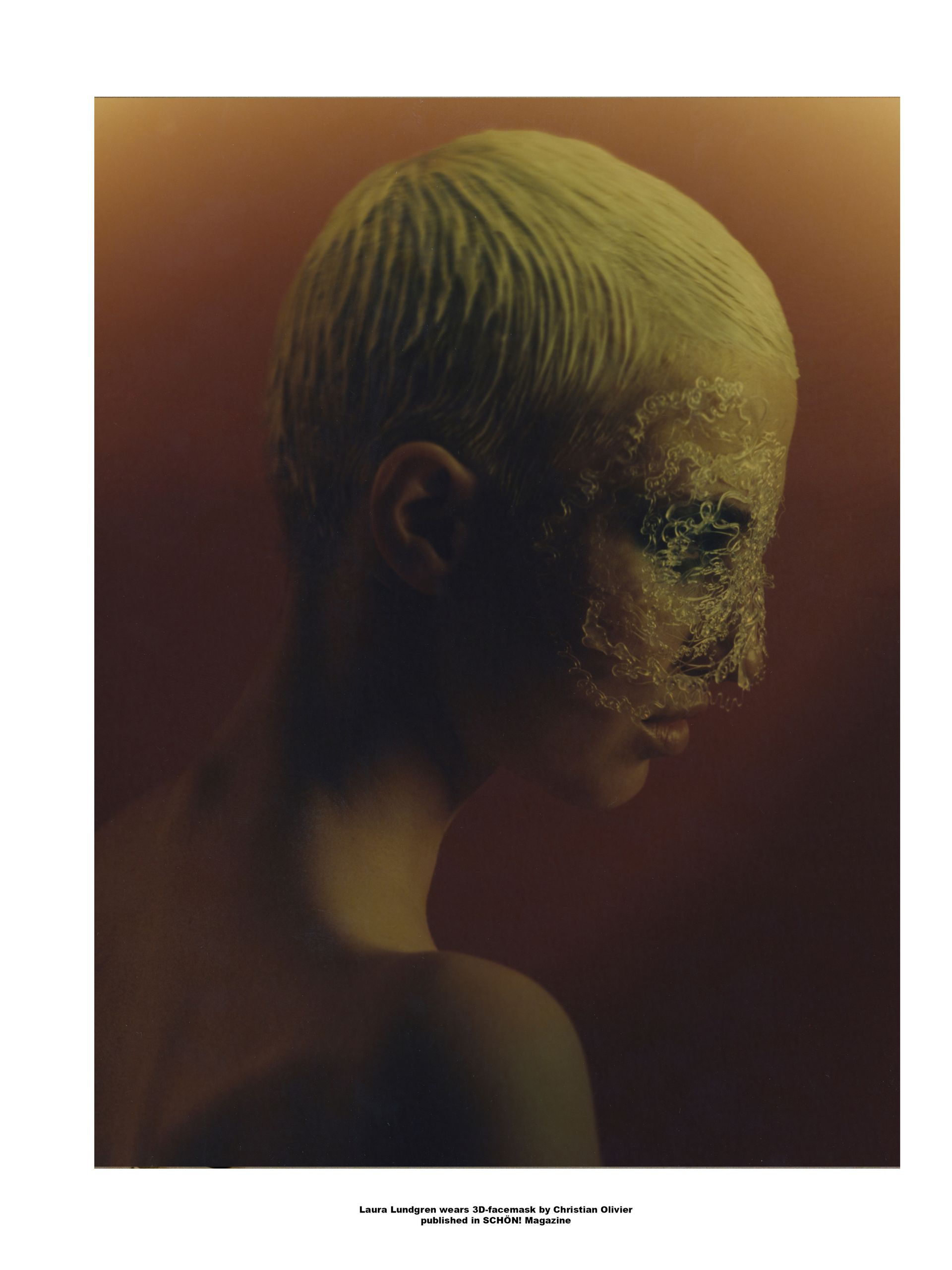 Frau mit kurzem Haar und teilweise mit einer strukturierten Substanz bedecktem Gesicht vor einem rotbraunen Hintergrund.