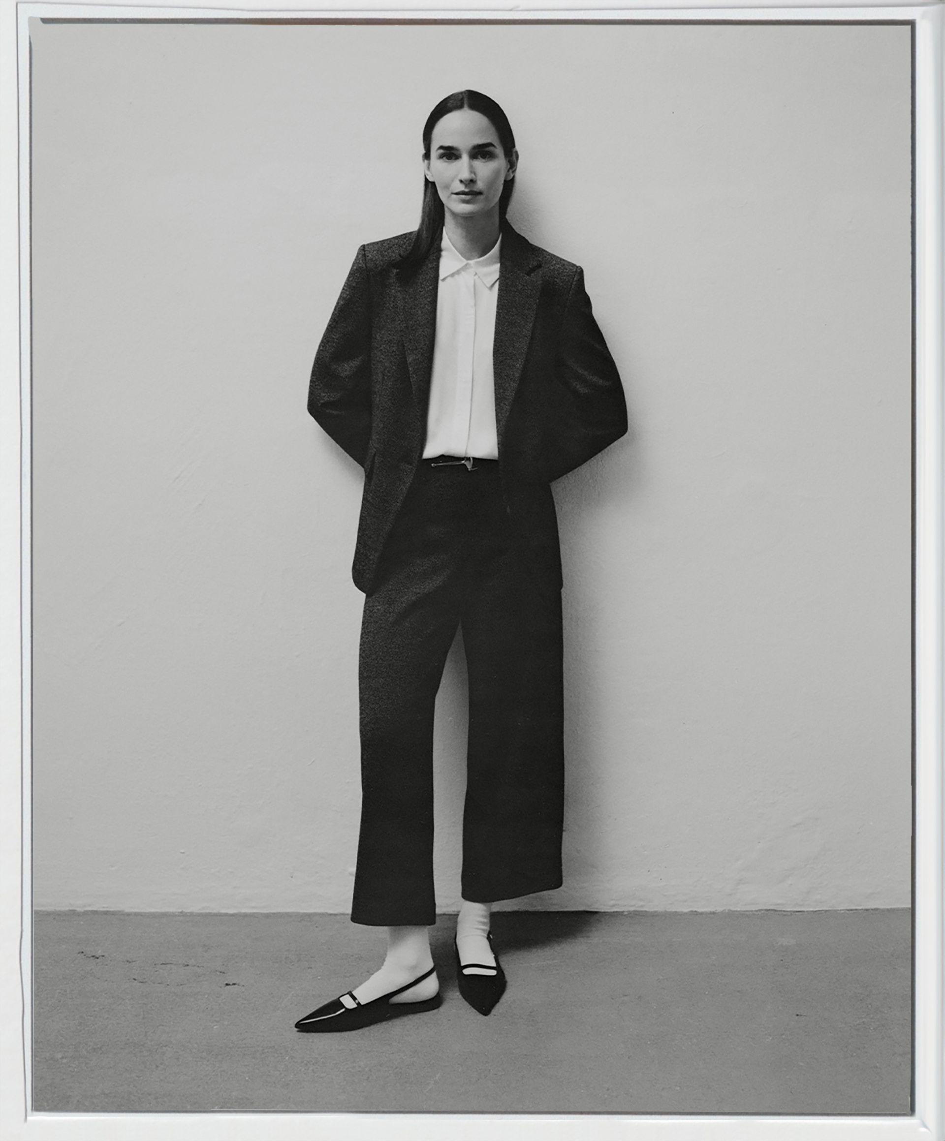 Frau in Blazer, Hose und flachen Schuhen steht vor einer schlichten Wand. Schwarz und Weiß.
