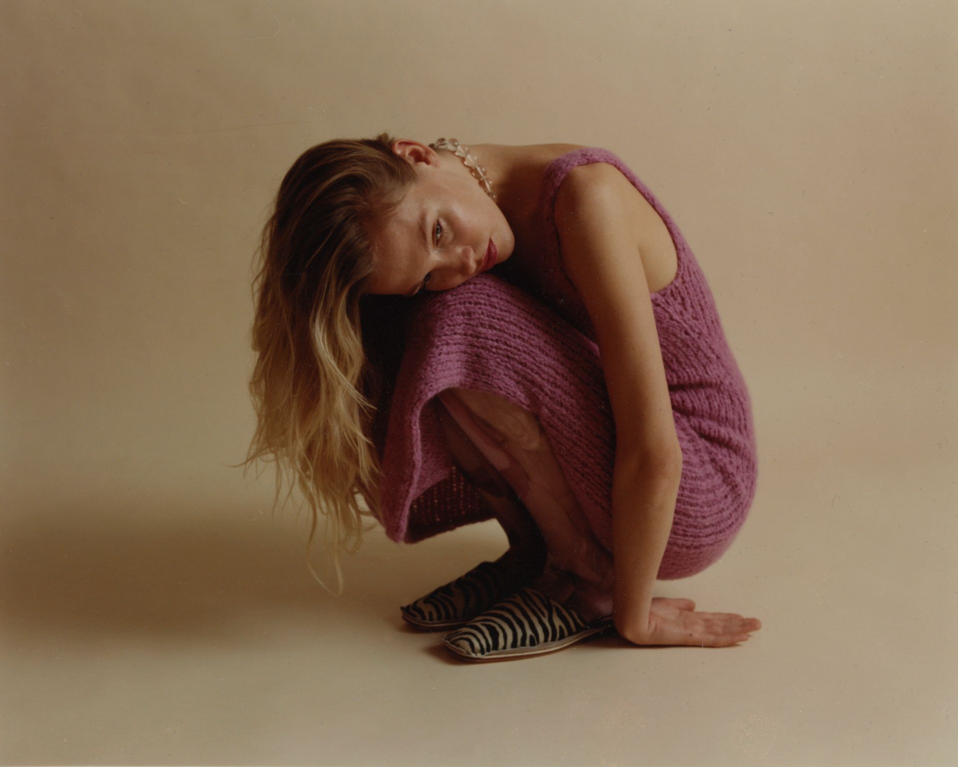 Frau im rosa Kleid, geduckt, Kopf auf den Knien ruhend; neutraler Hintergrund.