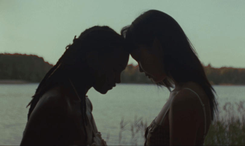 Zwei Frauen, Silhouetten, die Köpfe berühren sich. Eine mit Dreadlocks, eine mit langen Haaren, in der Nähe eines Sees.