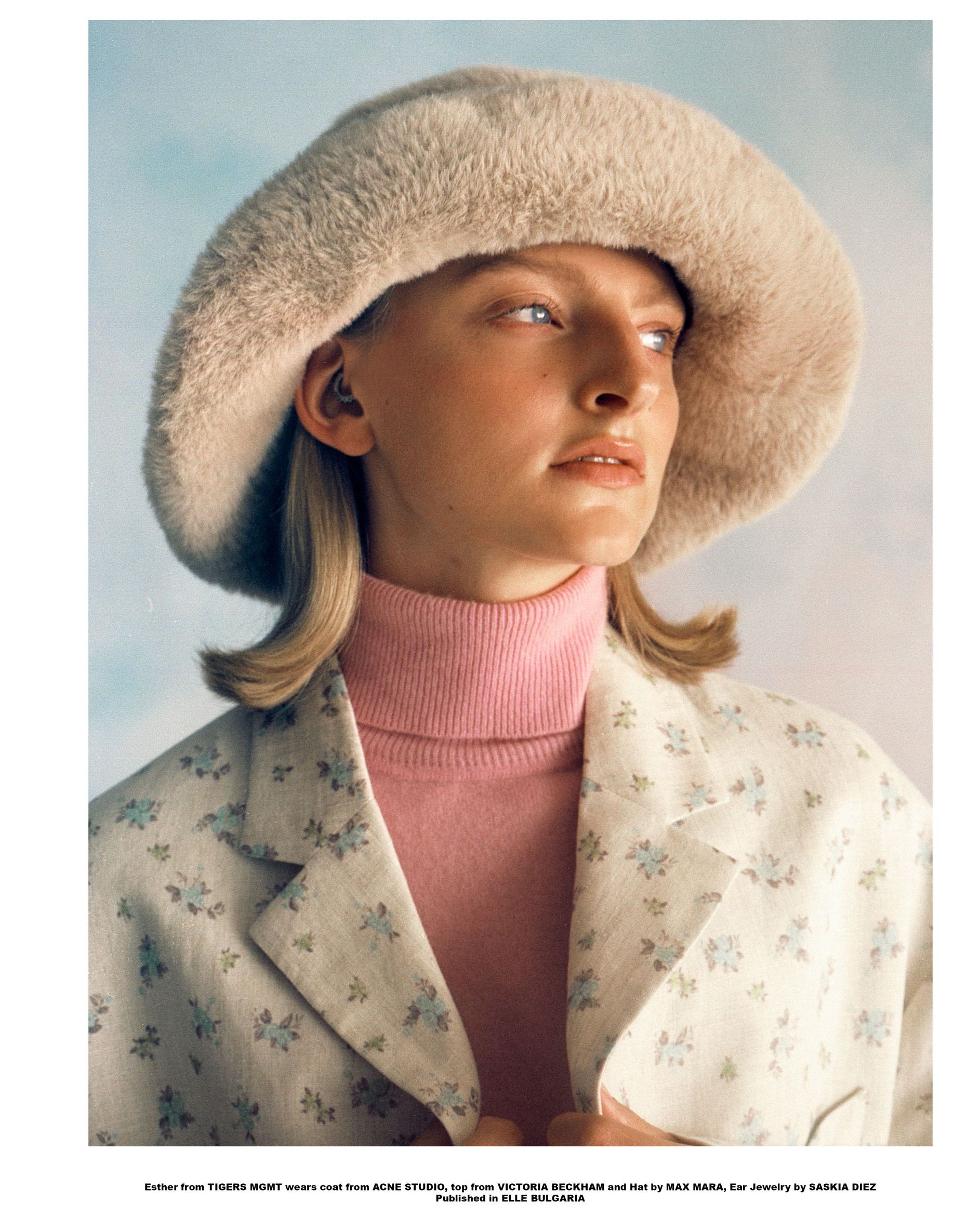 Frau mit beiger Pelzmütze, rosa Rollkragenpullover und geblümtem Blazer, Blick zur Seite.