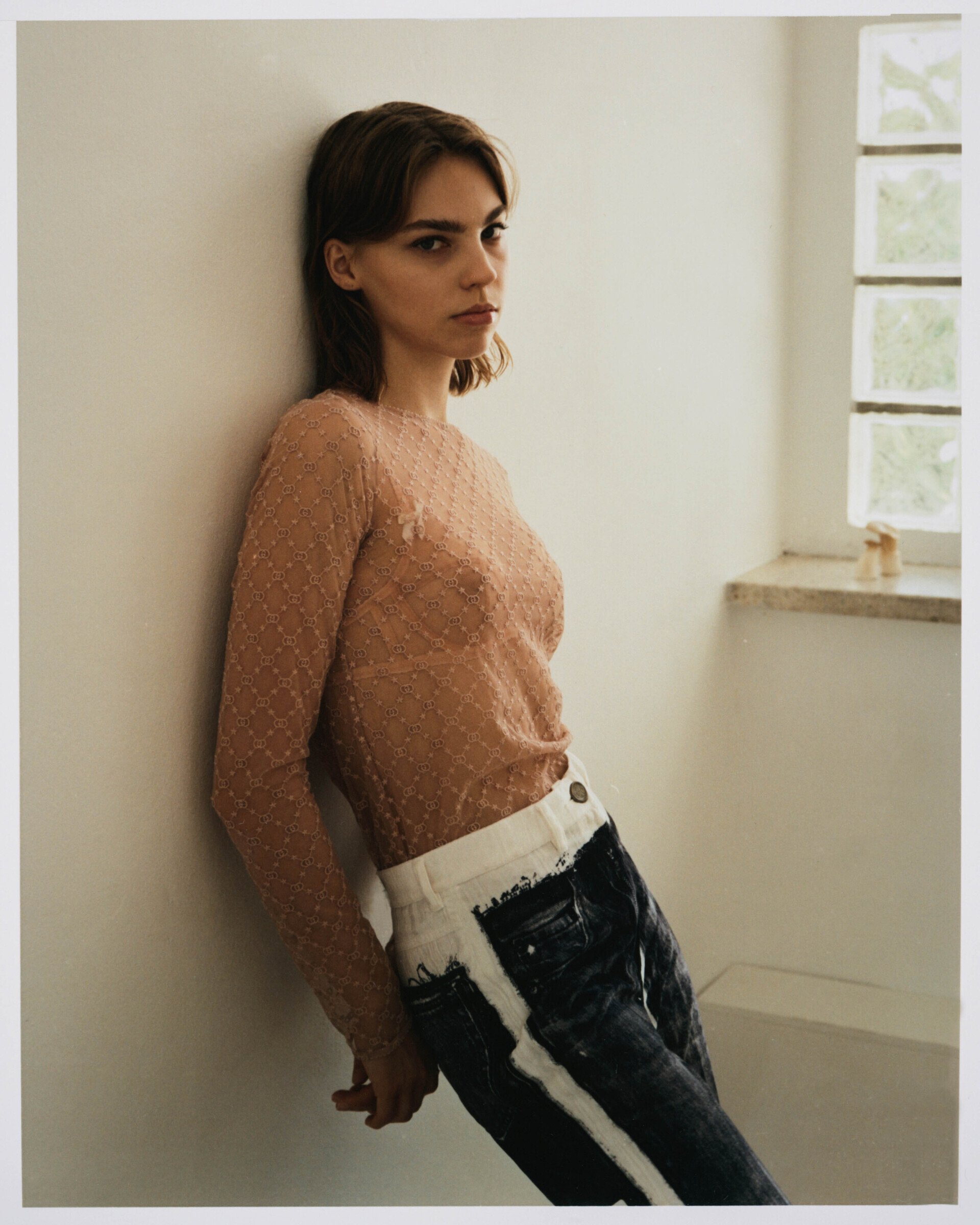 Frau in durchsichtigem rosa Top und Jeans lehnt an einer Wand neben einem Fenster.