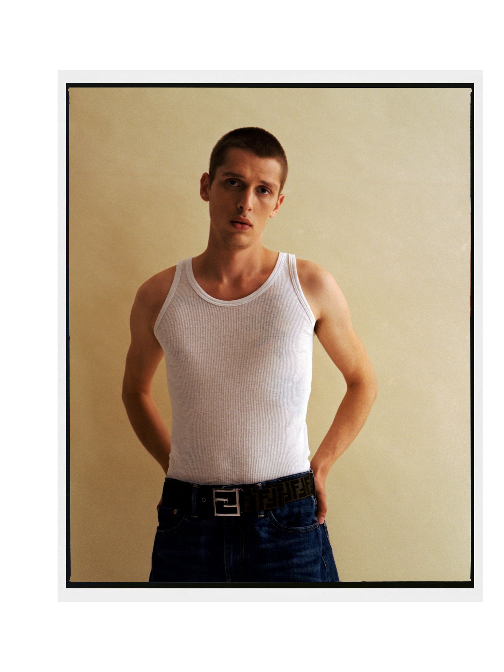 Mann in weißem Tanktop und Jeans posiert vor hellbraunem Hintergrund.