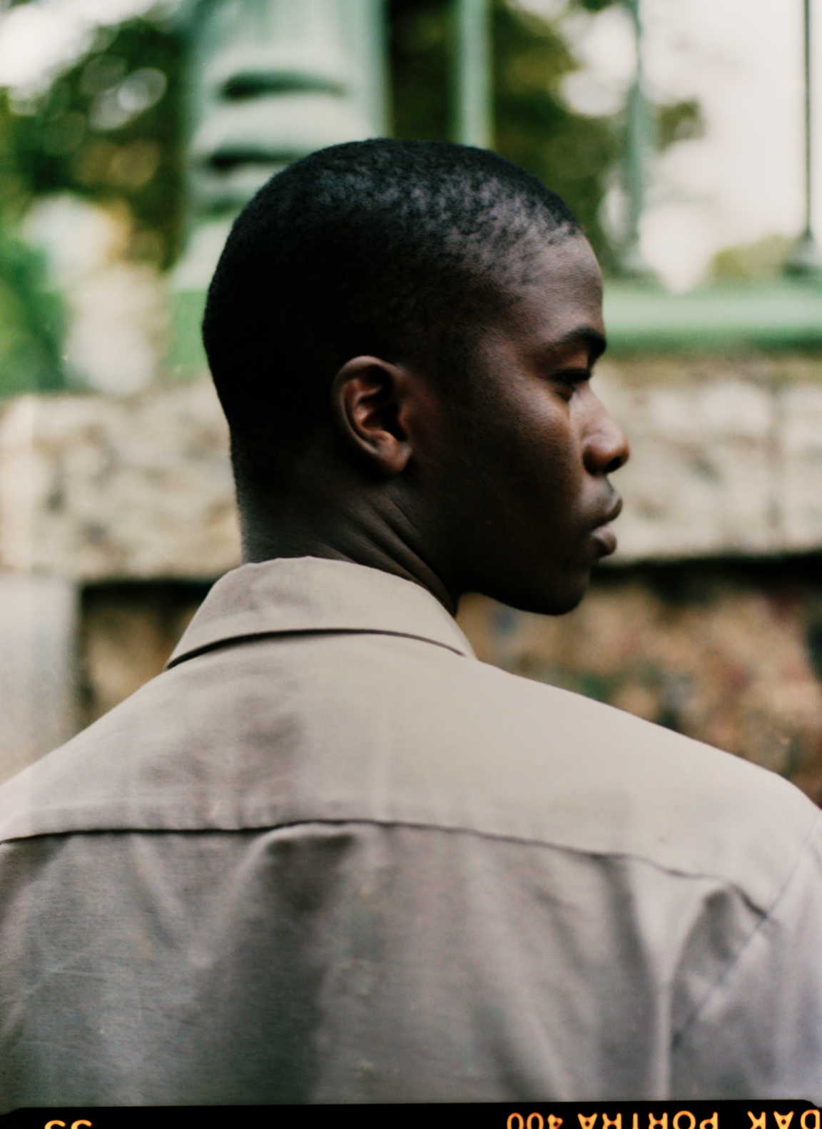 Junger Schwarzer in einem hellbraunen Hemd, zur Seite gedreht, im Freien.