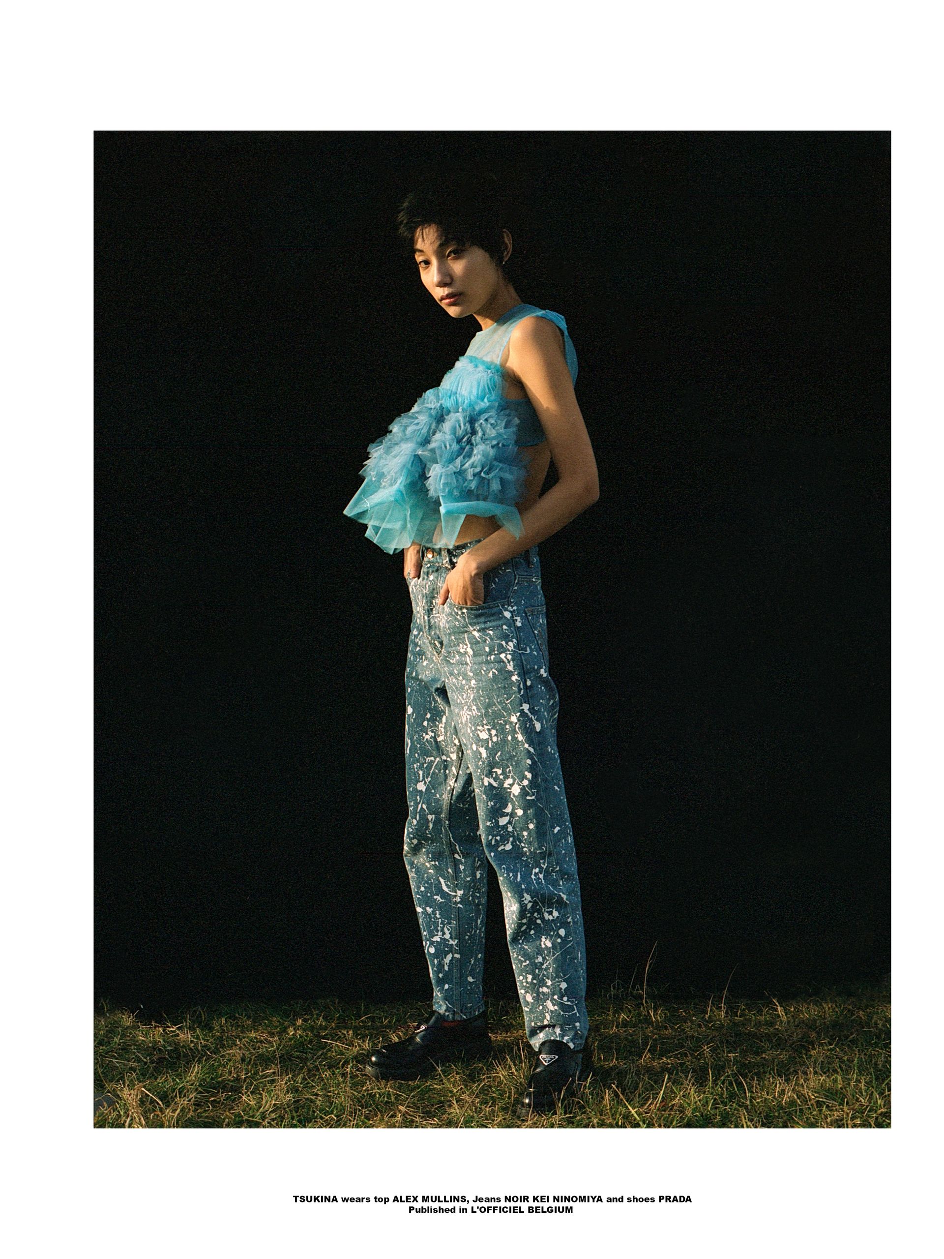 Frau in blauem Rüschentop und Paillettenjeans posiert auf Gras vor schwarzem Hintergrund.