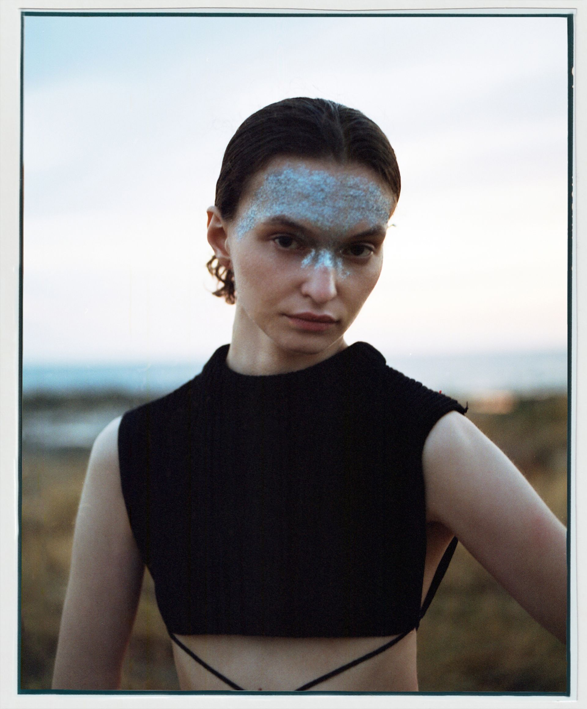 Frau mit Glitzer-Make-up im Gesicht, schwarzes Oberteil, blickt im Freien in der Nähe von Wasser in die Kamera.