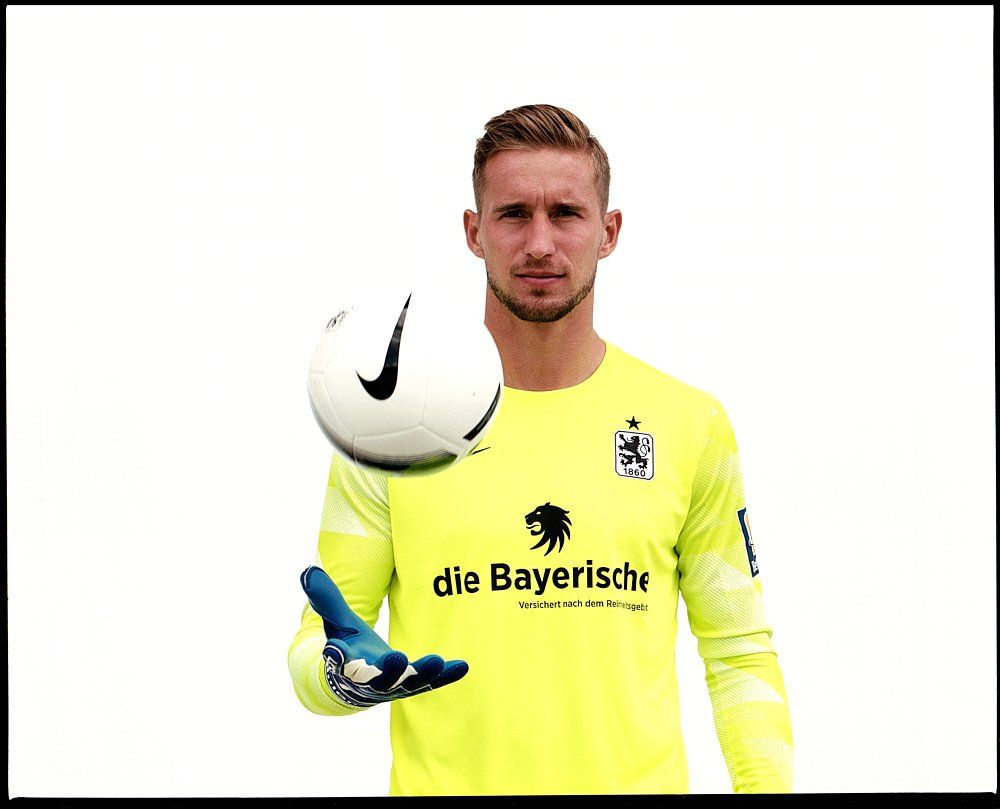 Torwart im hellgrünen Trikot fängt einen Fußball, weißer Hintergrund.